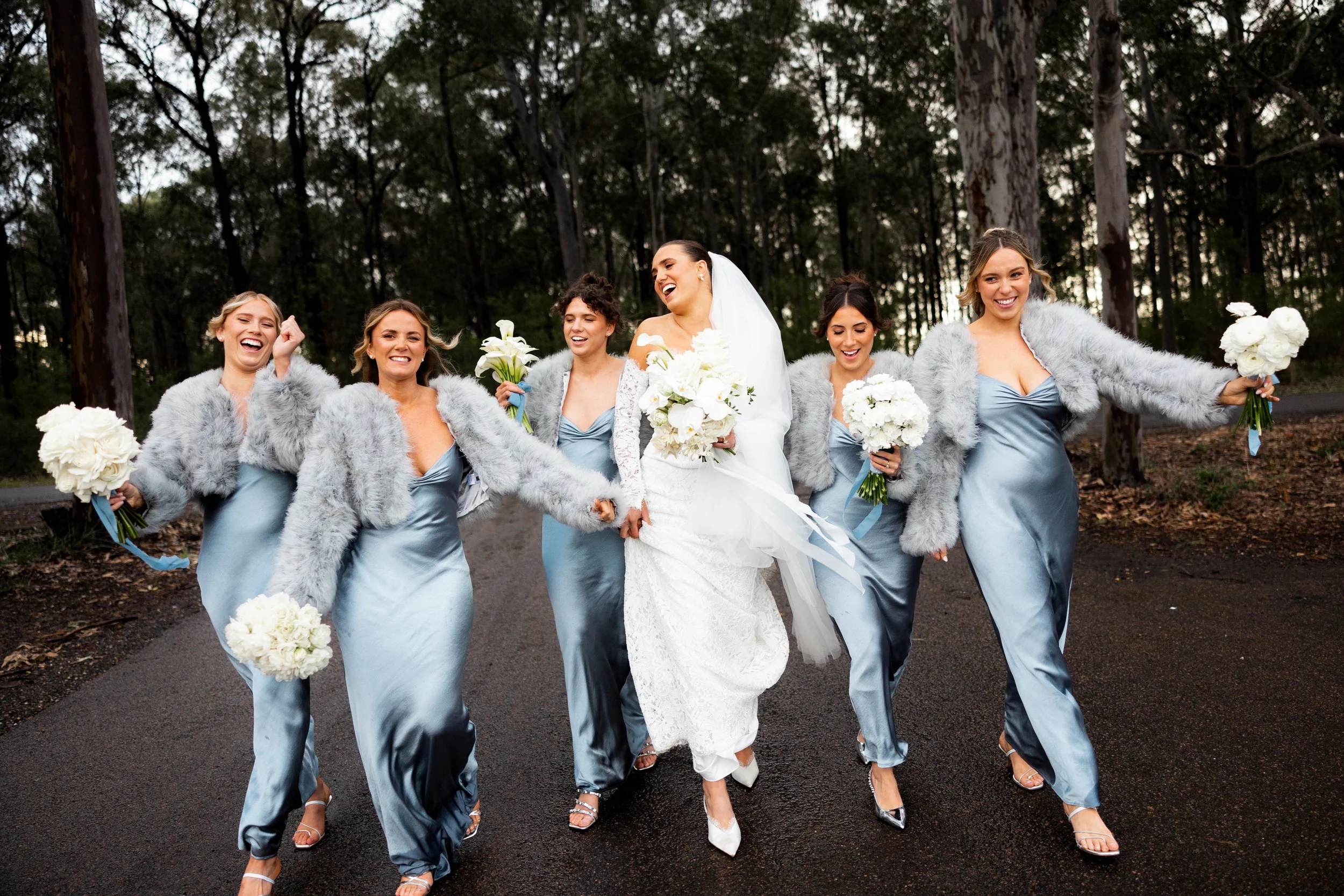 A bride with blonde hair in a white lace wedding dress, surrounded by five bridesmaids in dark green dresses, all holding colorful bouquets of flowers, standing outdoors near a white building.