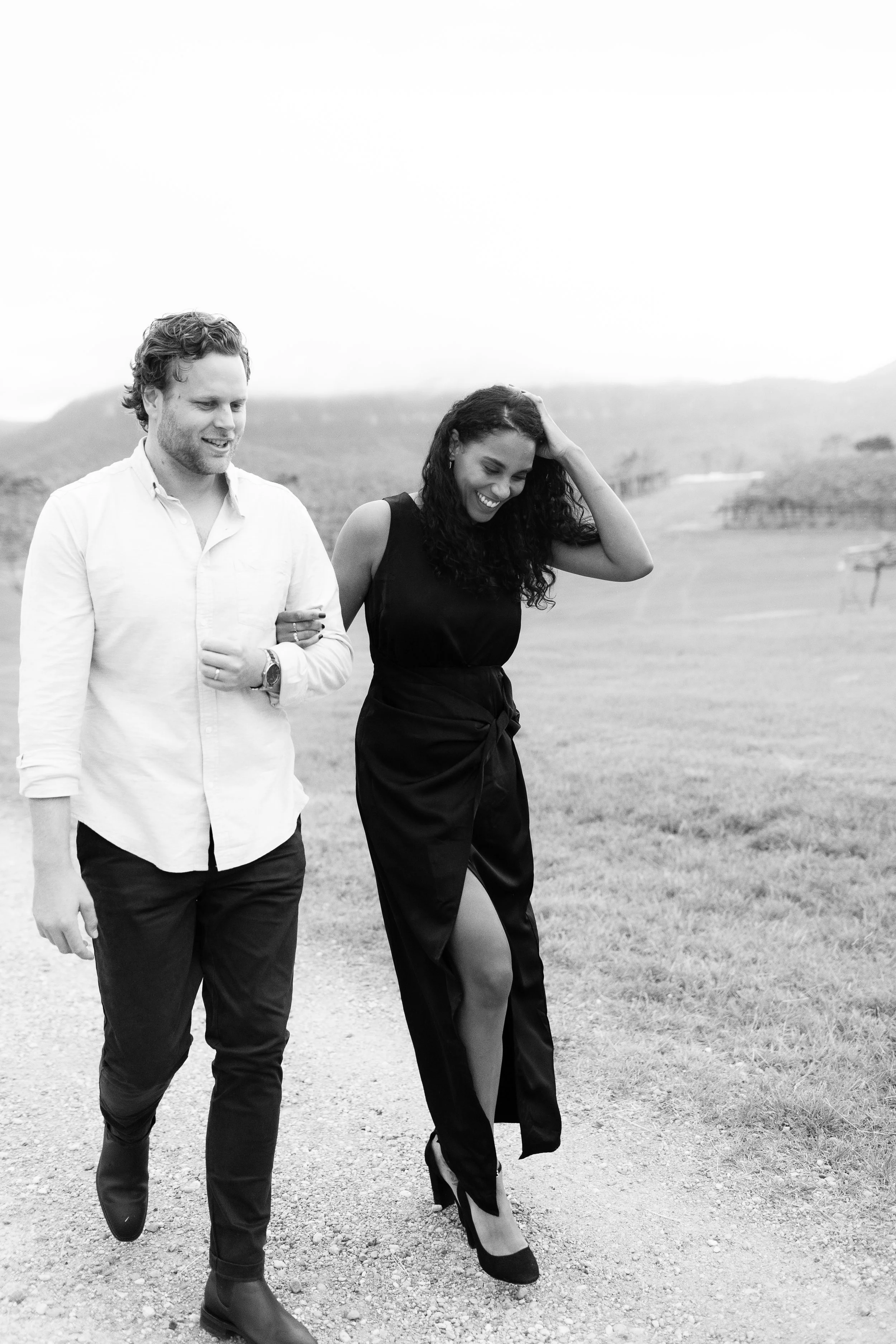 A man and woman walking outdoors on a gravel path, smiling and enjoying each other's company, with rolling hills and fields in the background, in black and white.