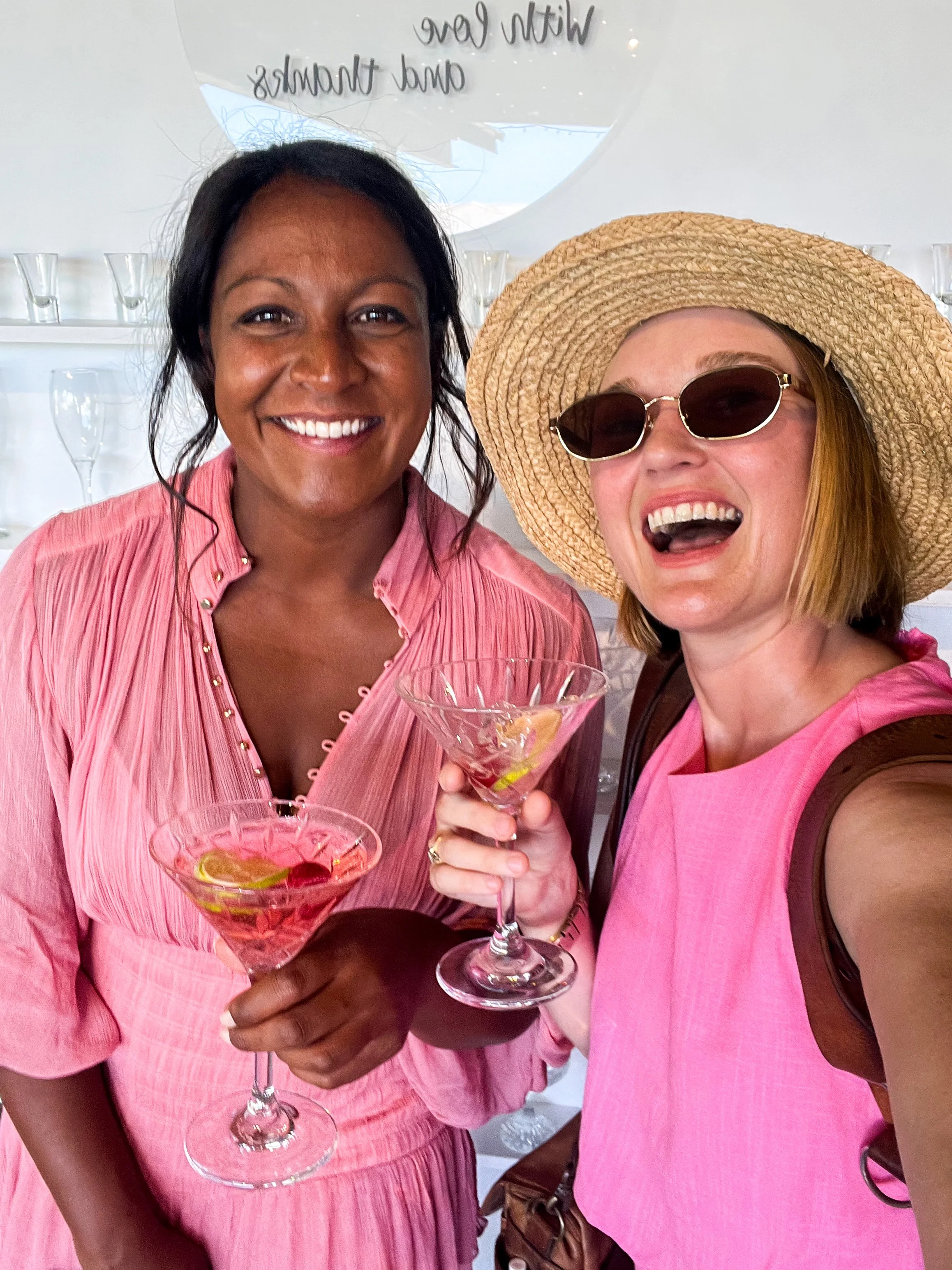 Two women smiling and holding cocktails, one wearing a pink dress and the other wearing a pink top with a large straw hat and sunglasses.