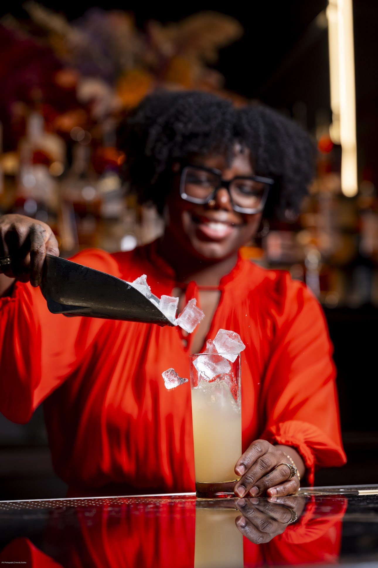 A woman with short curly hair, glasses, and a red blouse is smiling as she pours ice into a tall glass of yellowish cocktail at a bar.