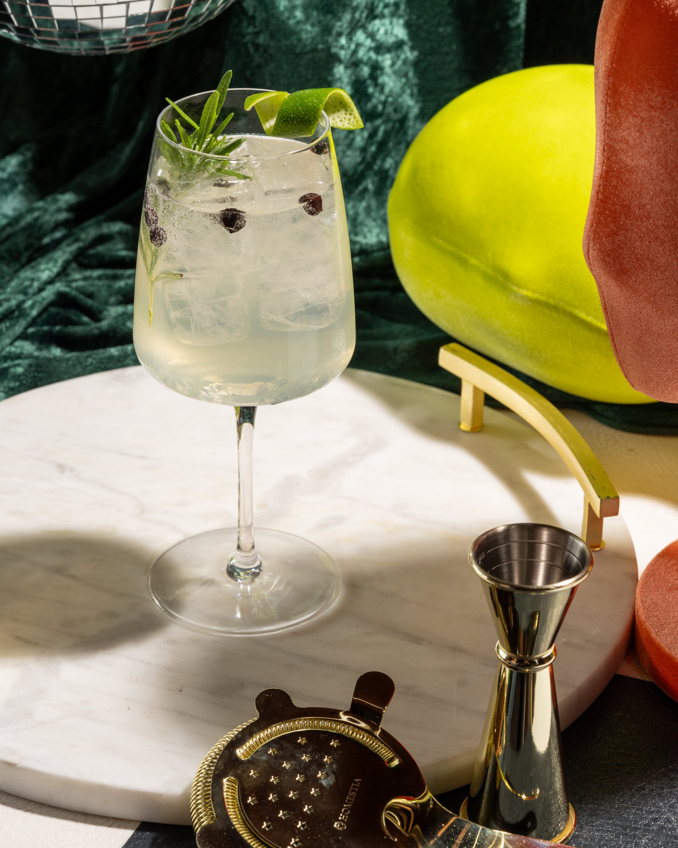 A glass of gin and tonic garnished with rosemary, lime, and juniper berries on a white marble surface with a green velvet backdrop, a yellow decorative object, an orange fabric, and bar tools including a jigger and a round gold coaster.