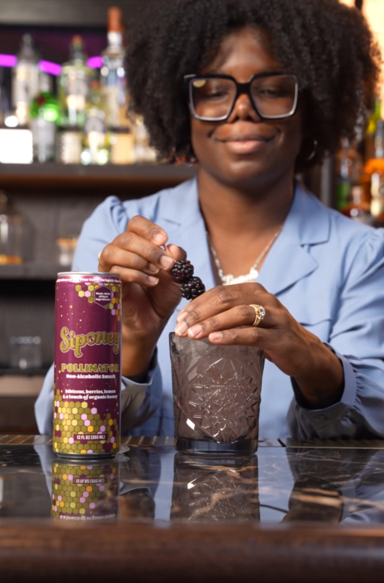 A woman with glasses and curly hair adds blackberries to a glass at a bar, with a can of Siponee pollinator organic berry and lemon drink on the table in front of her.