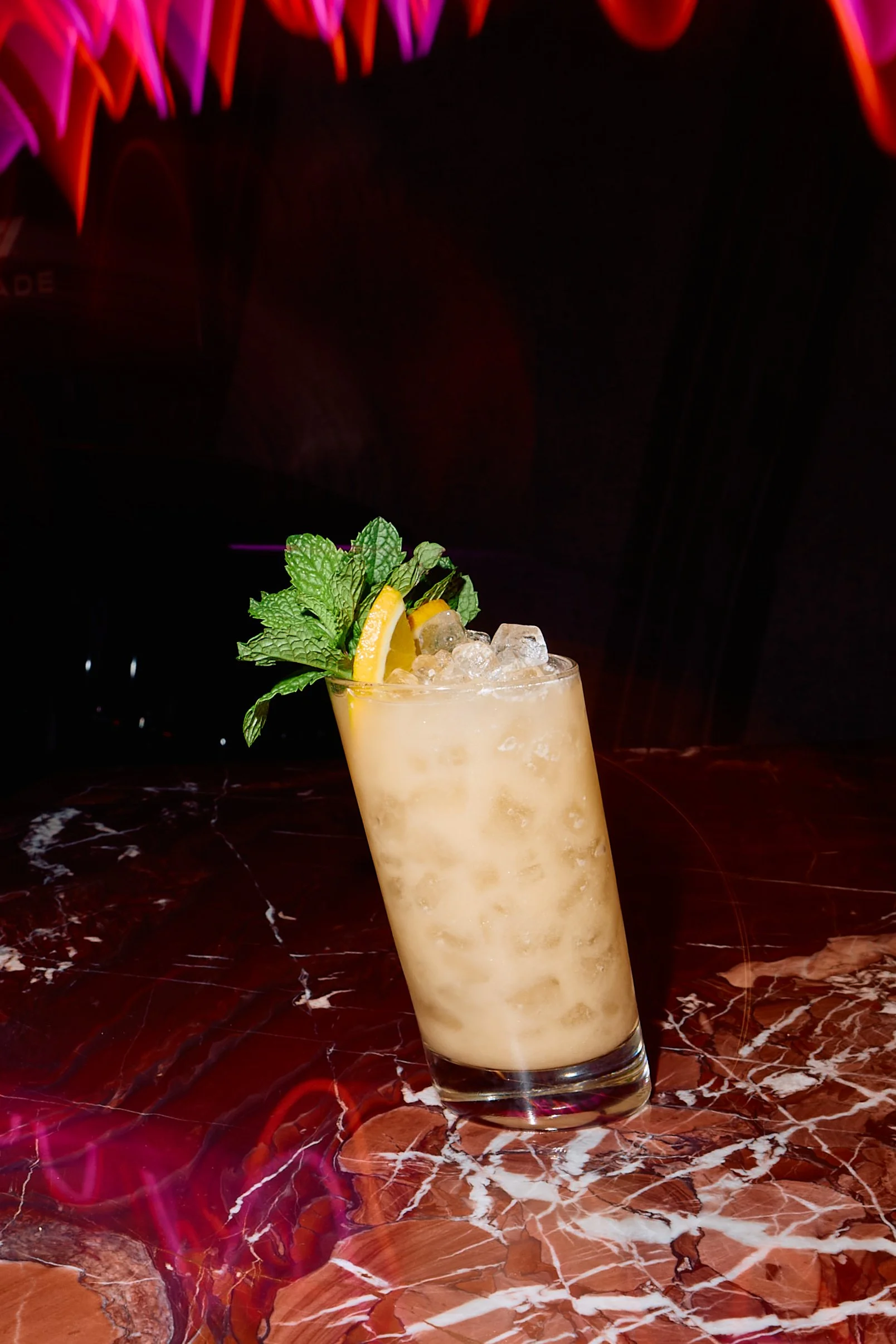 A highball glass filled with a creamy, light-colored cocktail, garnished with lemon slices and fresh mint leaves, on a dark marble surface.