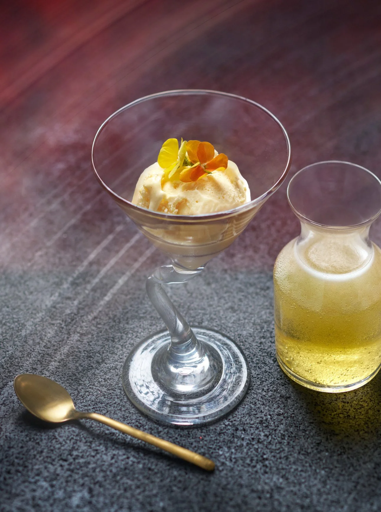 A glass of vanilla ice cream garnished with yellow and orange edible flowers, accompanied by a glass pitcher of a light yellow beverage, and a small gold spoon on a dark textured surface.