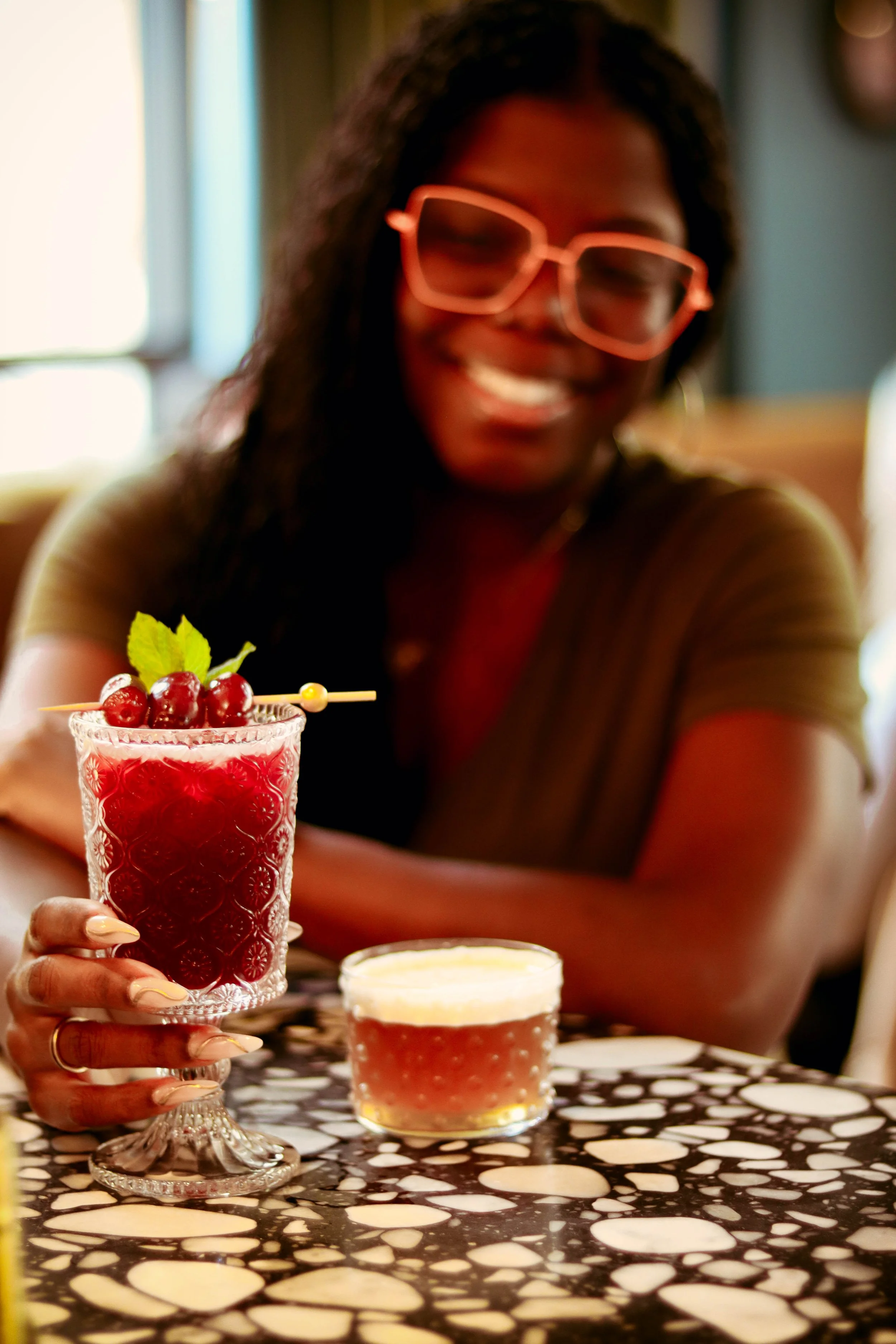 A woman with glasses smiling and holding a glass with red beverage and garnished with red grapes and mint leaves, sitting at a table with a patterned black and white surface, with a small bowl of red drink in front of her. The background is softly li