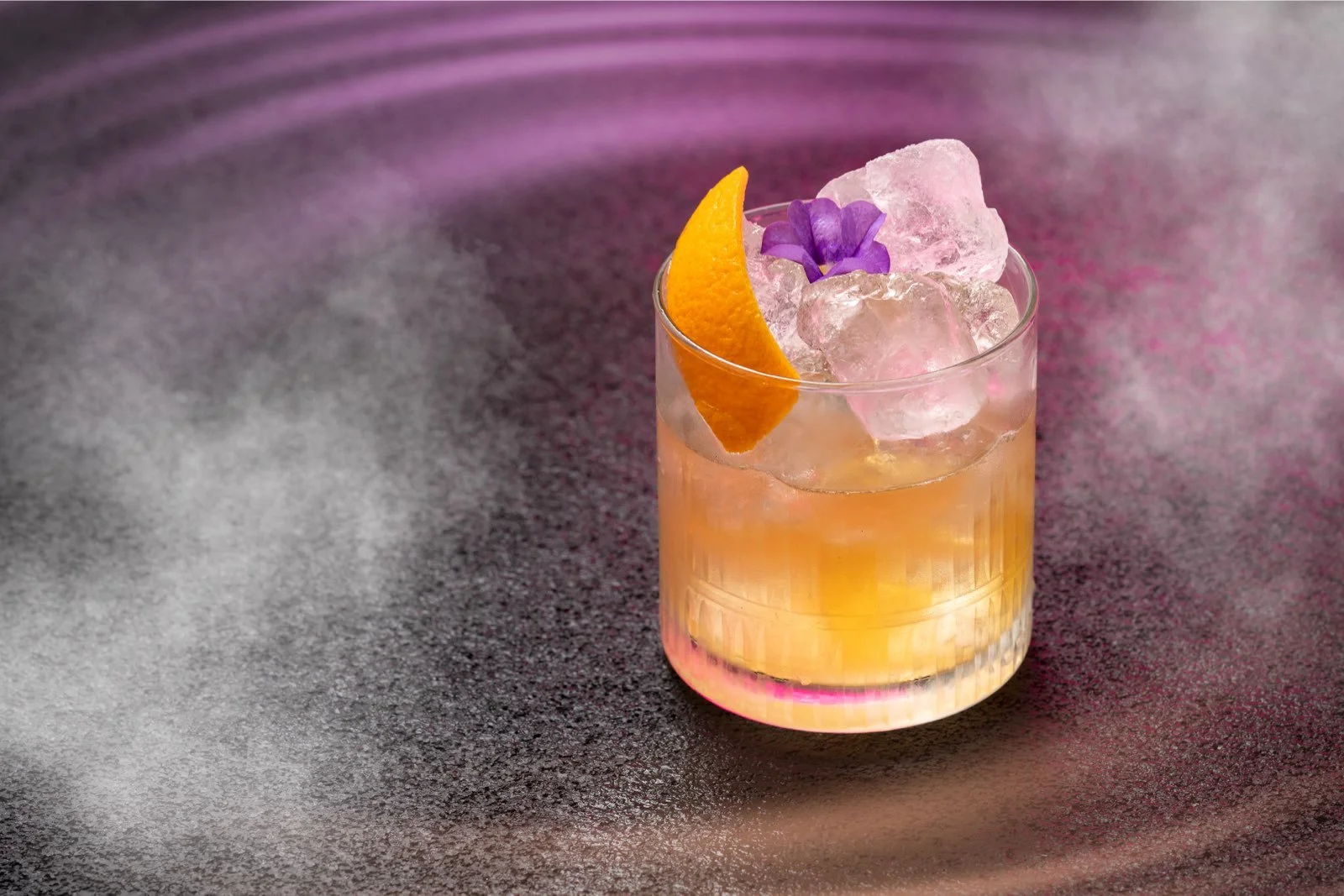 A cocktail in a short glass with ice, garnished with an orange slice and a purple flower, against a textured purple and pink background.