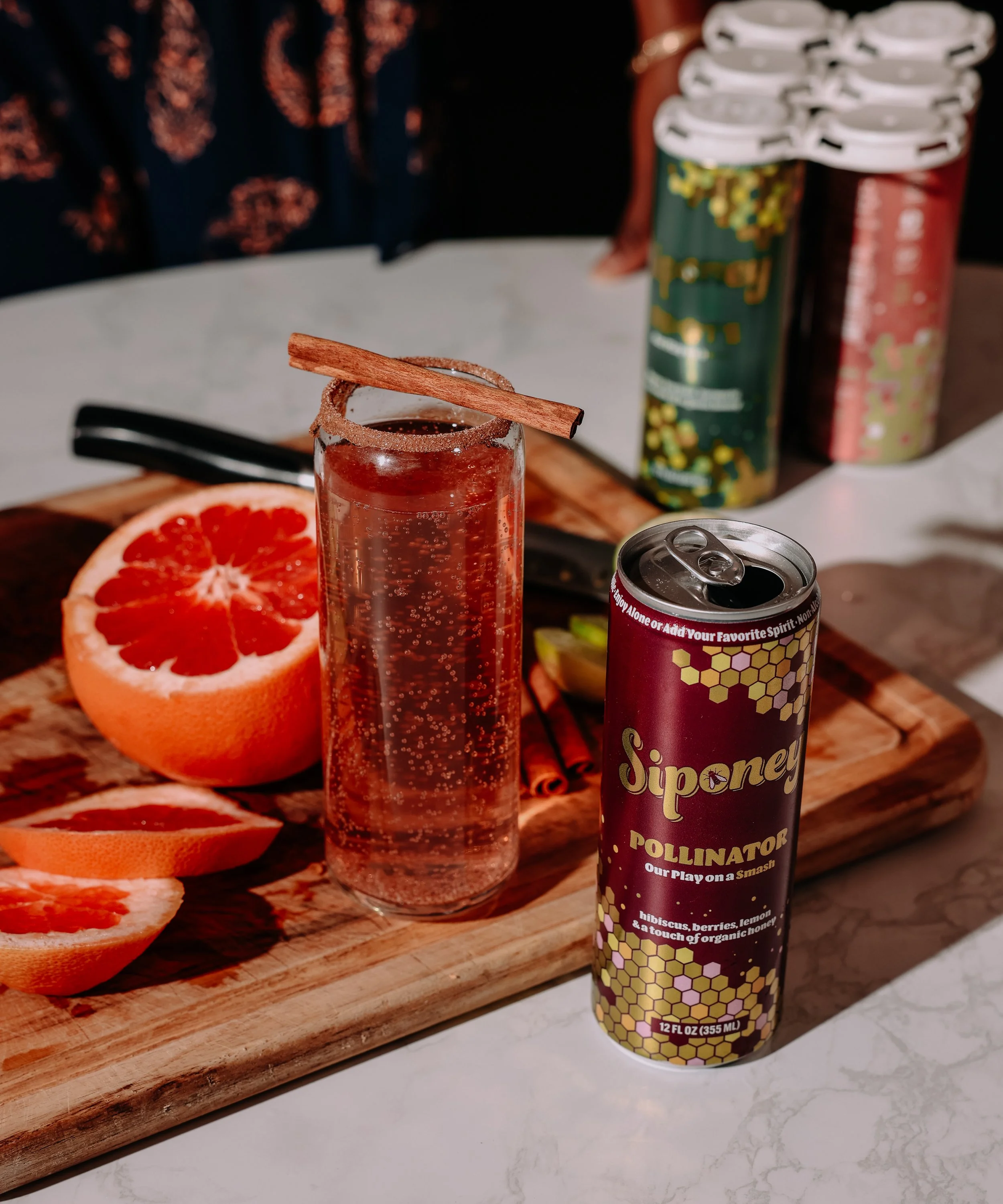A glass of sparkling beverage with a cinnamon stick garnish on a marble table, alongside a halved grapefruit and a can of Siponey Pollinator soda, with additional cans in the background.
