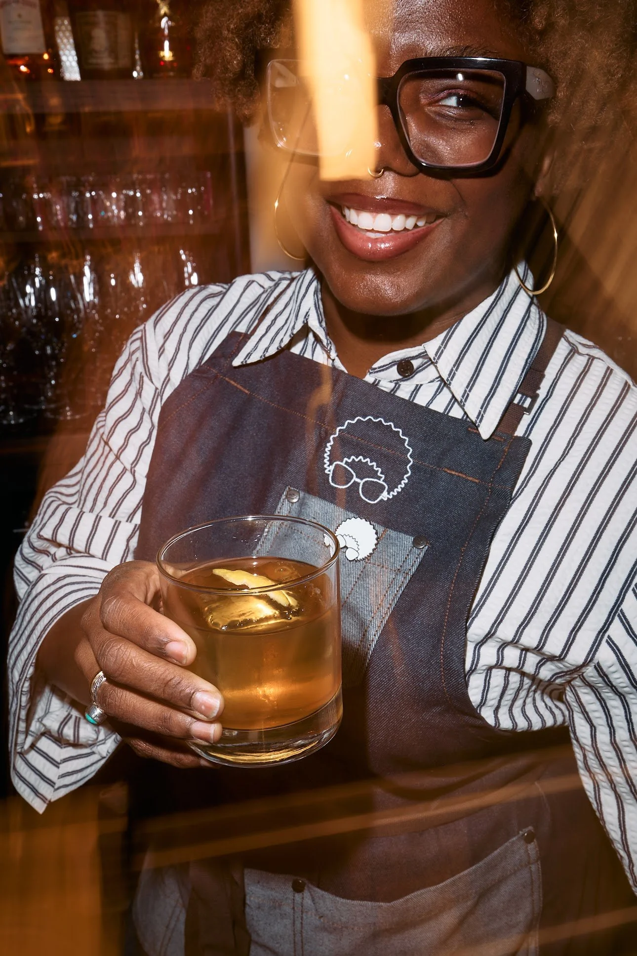 Woman wearing glasses and an apron holding a glass of whiskey or similar amber-colored drink with ice, smiling in a bar or restaurant setting.