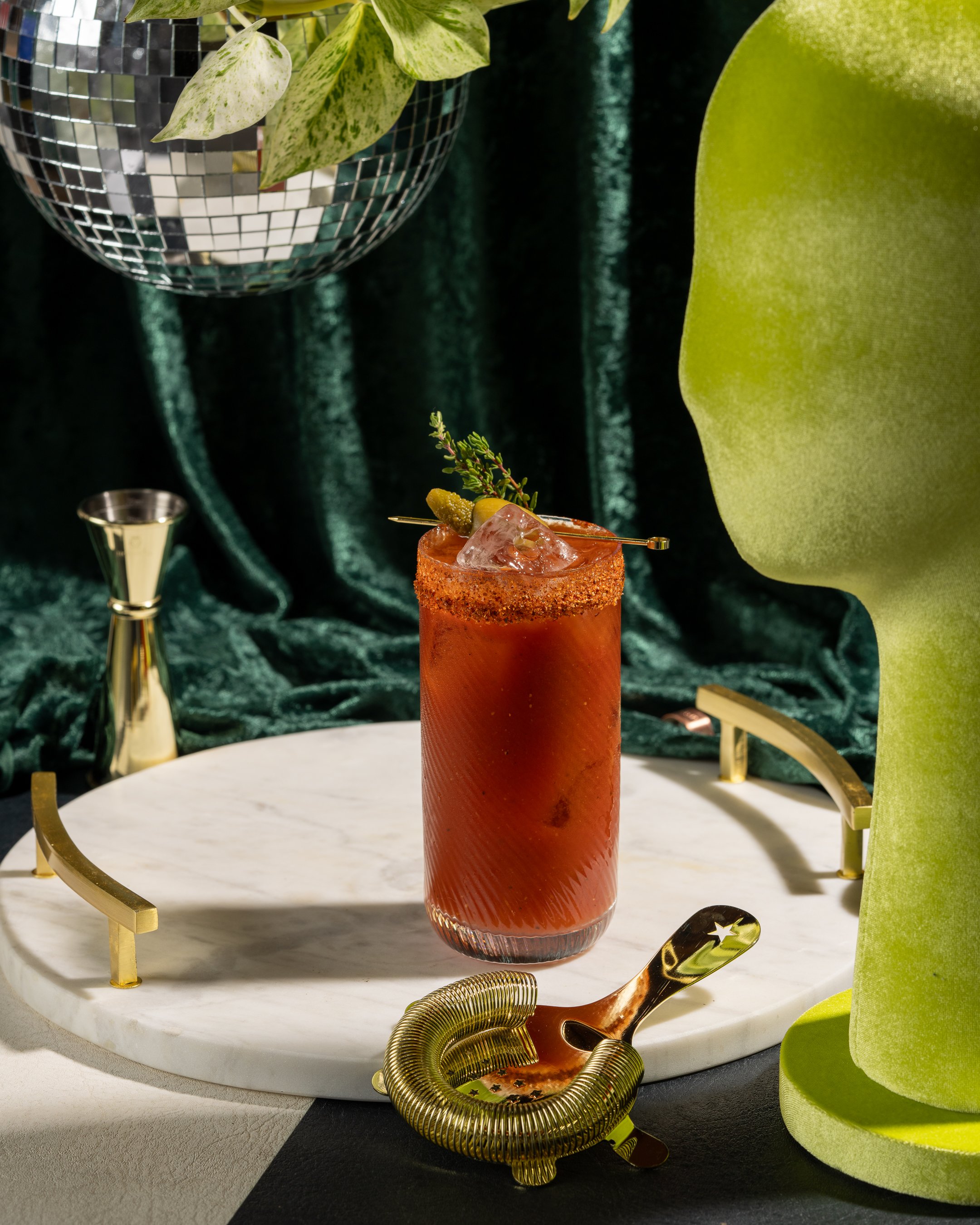 A tall red cocktail with ice and garnishes on a white marble tray, surrounded by various decorative items including a disco ball, green velvet fabric, and gold accents.