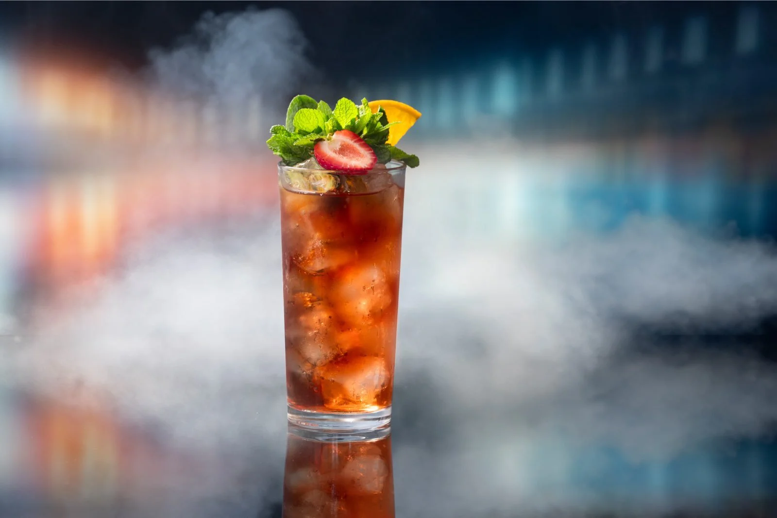 A tall glass of iced tea garnished with a strawberry, mint leaves, and a lemon wedge, set against a blurred modern bar background.