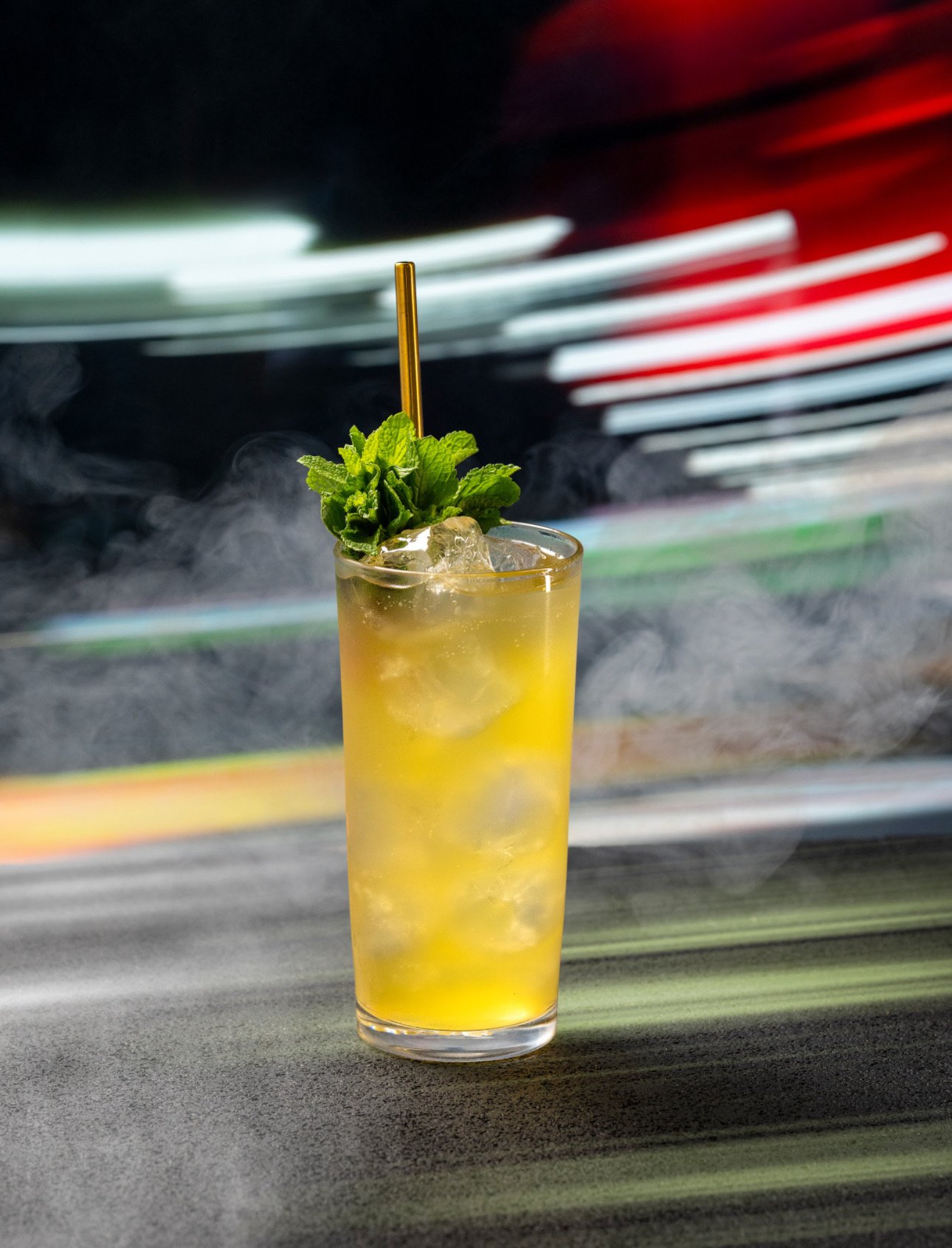 Tall glass of yellow cocktail with ice, garnished with fresh mint leaves and a gold straw, on a dark surface with blurred colorful lights in the background.