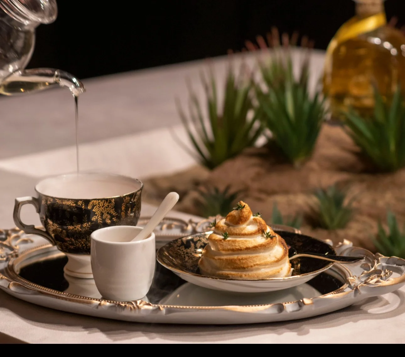 A dessert with a swirl of meringue topped with small pastry dollops on a black and gold ornate plate, next to a black and gold teacup, a white creamer with a spoon, and a glass teapot pouring liquid, all on a silver tray with greenery and bottles in 