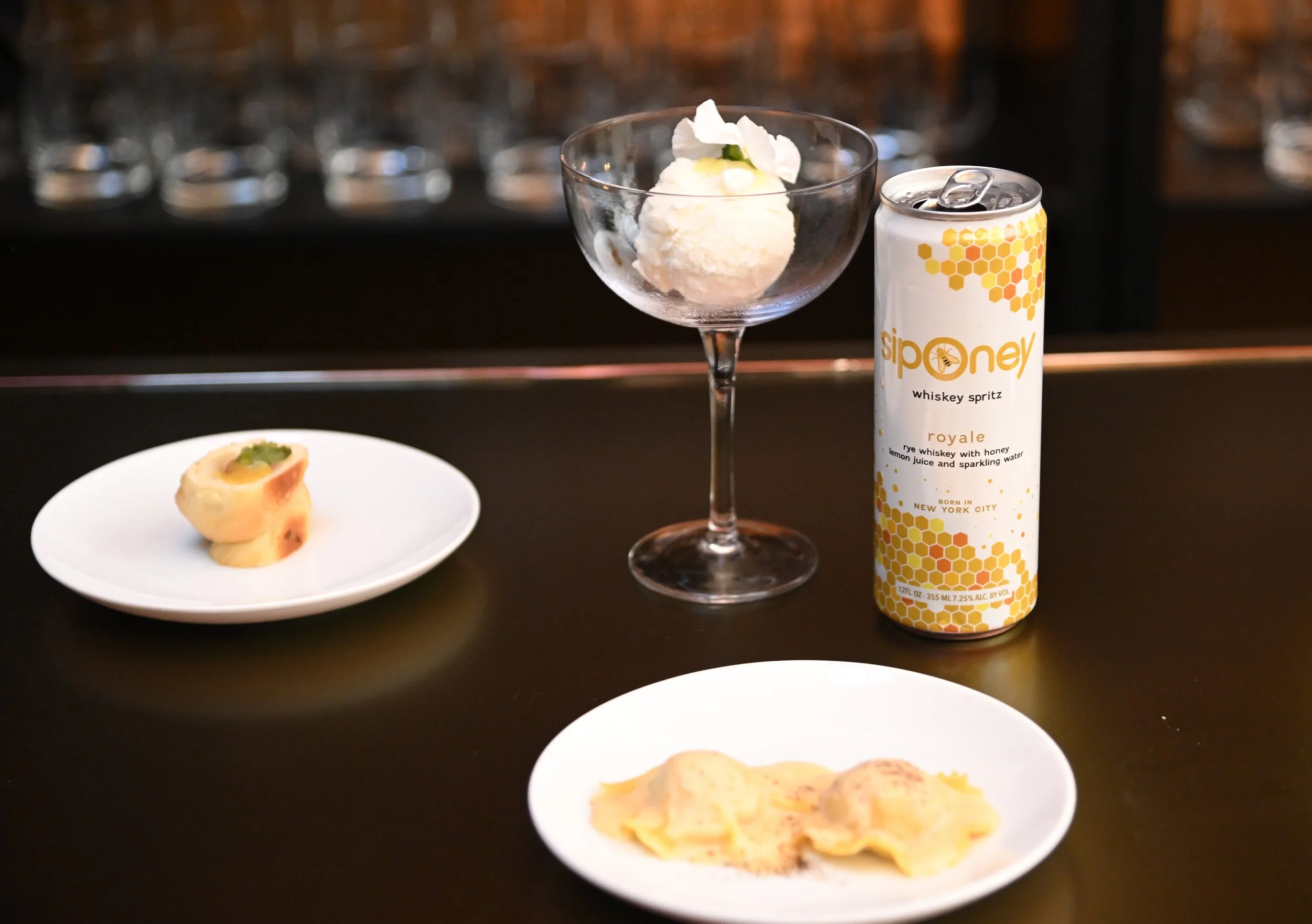 A scoop of vanilla ice cream garnished with a flower in a glass dessert bowl, a canned whiskey spritz drink, a small plate of bite-sized appetizers, and a white plate with a partially eaten piece of food on a dark bar or table surface.