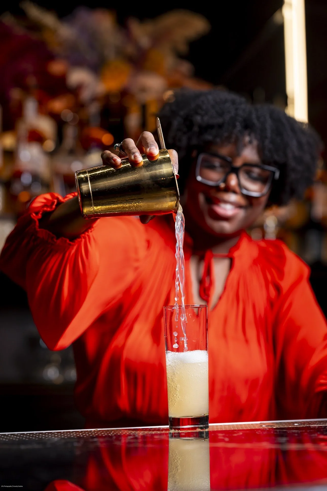 Person pouring a drink into a glass at a bar, wearing an orange top and glasses, with a blurred background of bottles.