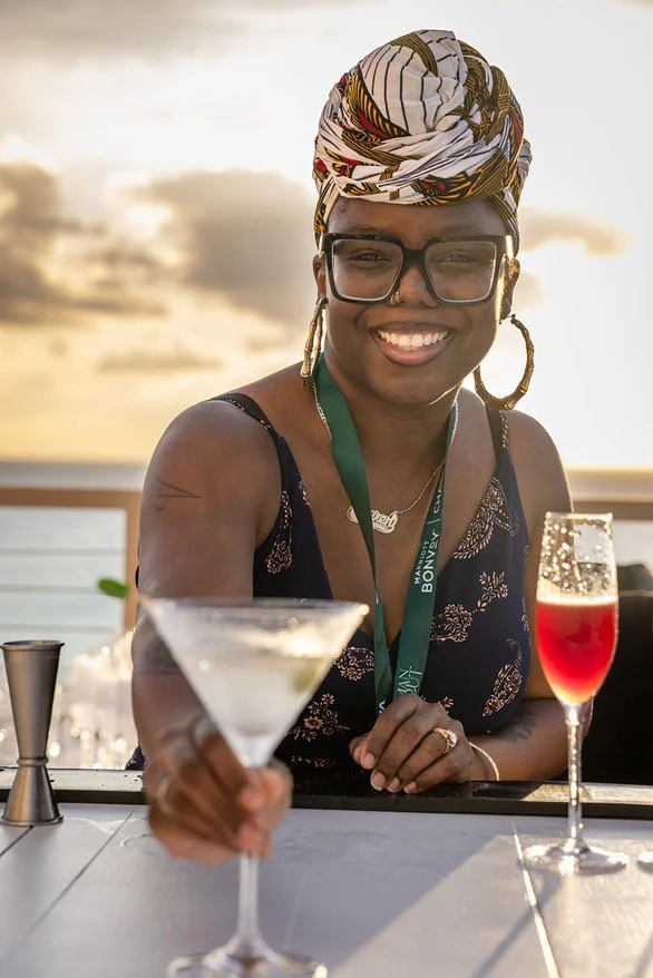 A smiling woman wearing glasses and a colorful headwrap, holding a cocktail, standing outdoors at sunset with a sunset sky and ocean in the background.