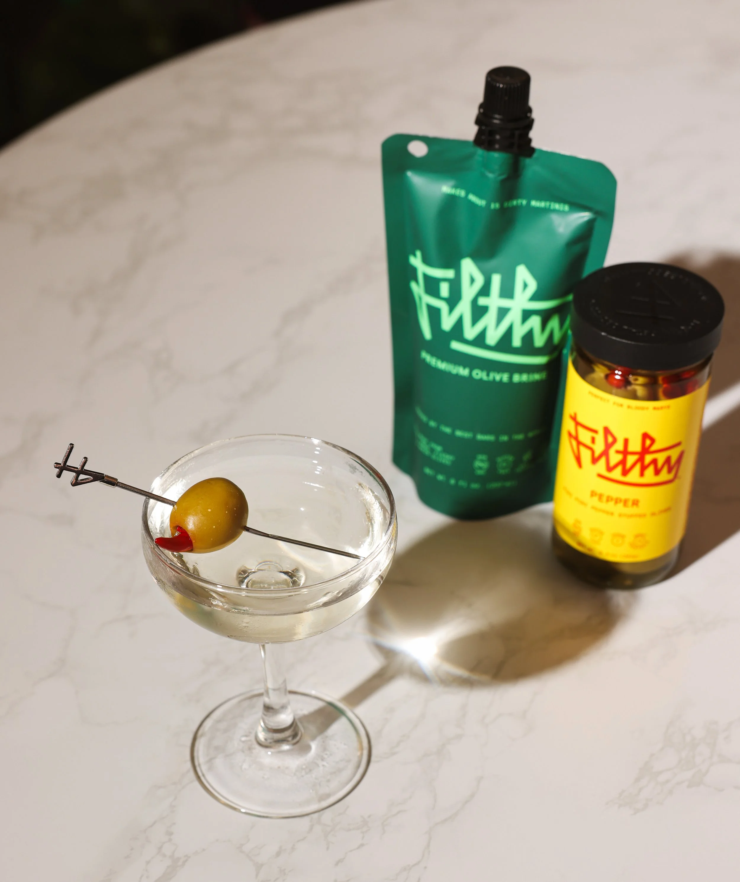 A cocktail glass with an olive, a green squeeze bottle, and a yellow spice jar on a white marble table.