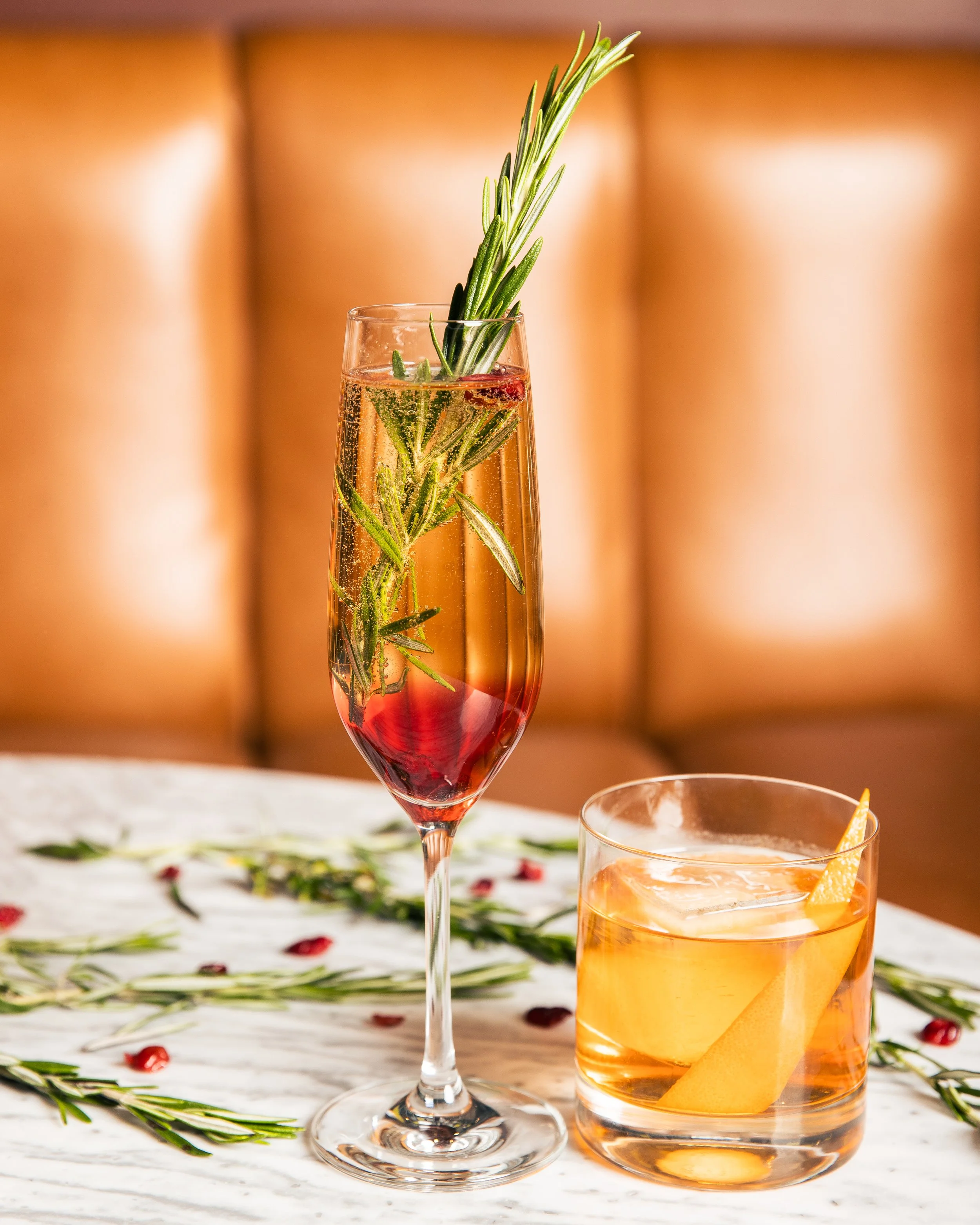 A glass of sparkling rosé with rosemary sprigs and a slice of lemon, garnished with a sprig of rosemary, on a white marble table with scattered rosemary and pomegranate seeds, behind a wooden booth.