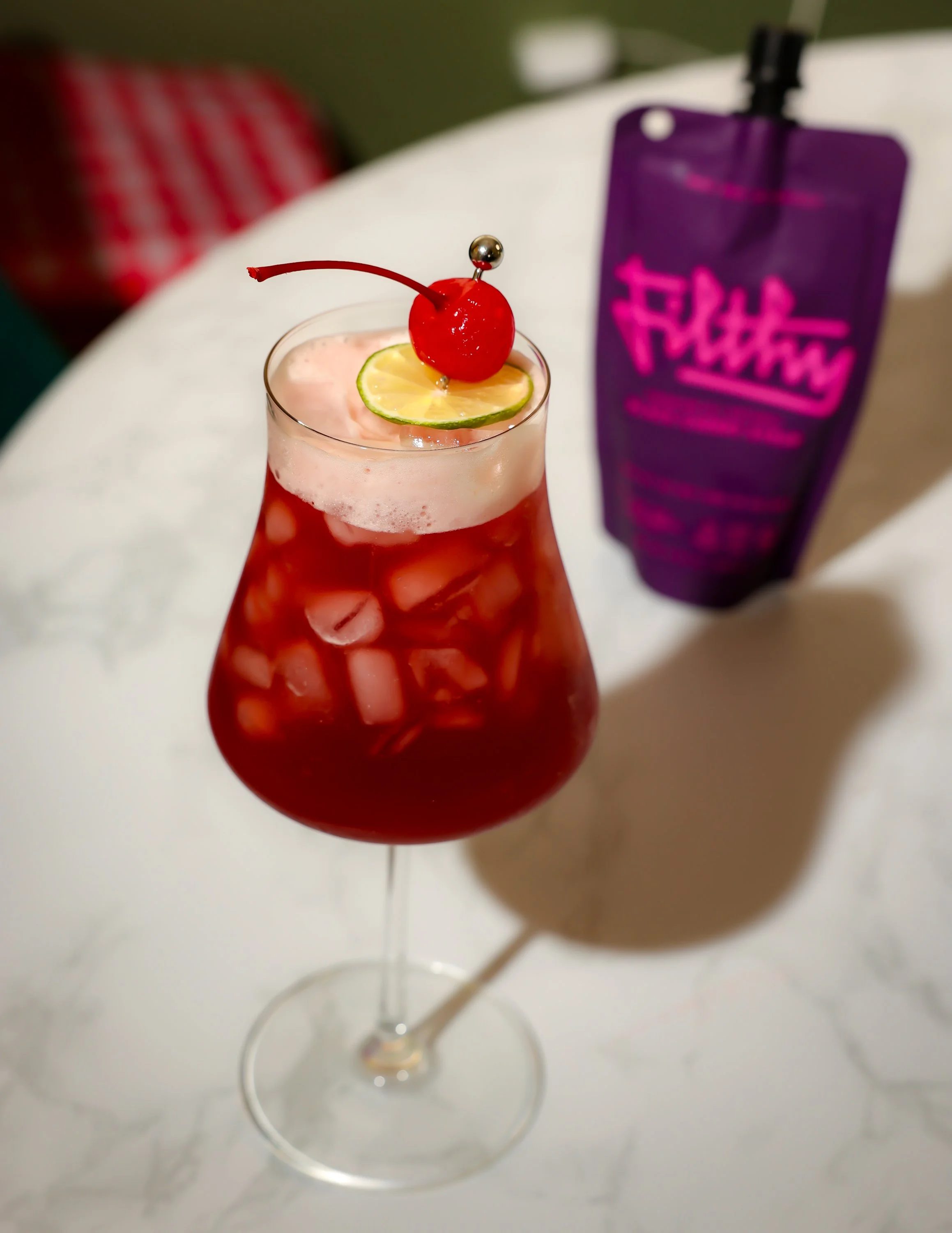 A cocktail glass filled with red-colored drink and ice, garnished with a lime slice and a maraschino cherry on top. There is a purple bottle in the background.