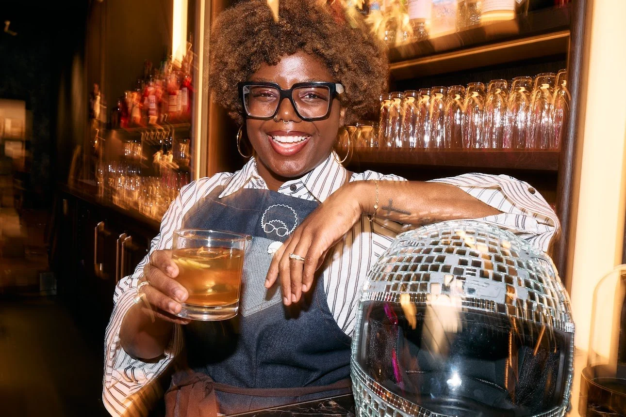 A smiling woman with glasses and a nose ring, wearing a striped shirt and apron, holding a glass of whiskey in a bar or restaurant setting.
