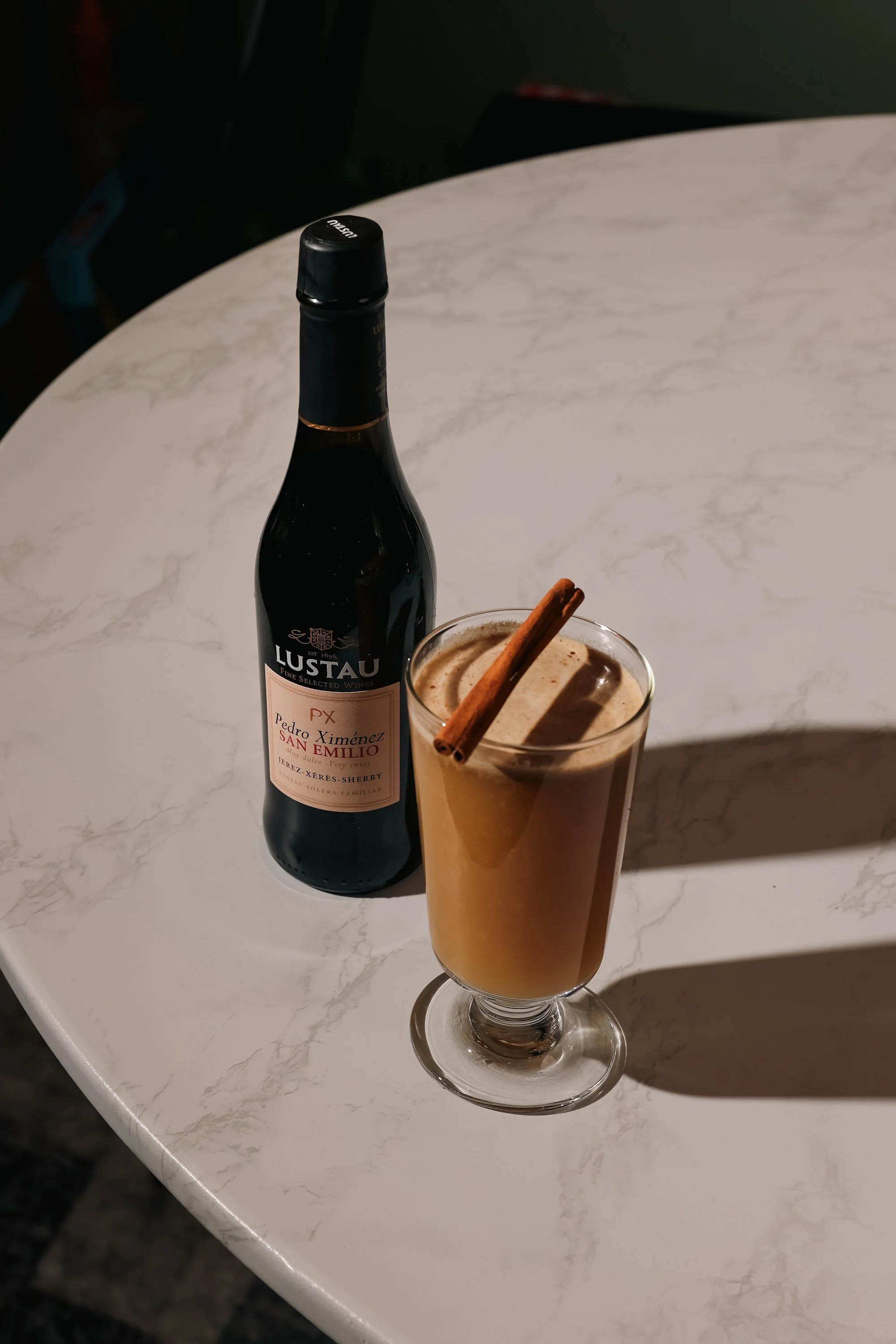 A bottle of Lustau Pedro Ximénez San Emilio sherry and a tall glass of creamy beverage garnished with a cinnamon stick on a white marble table.