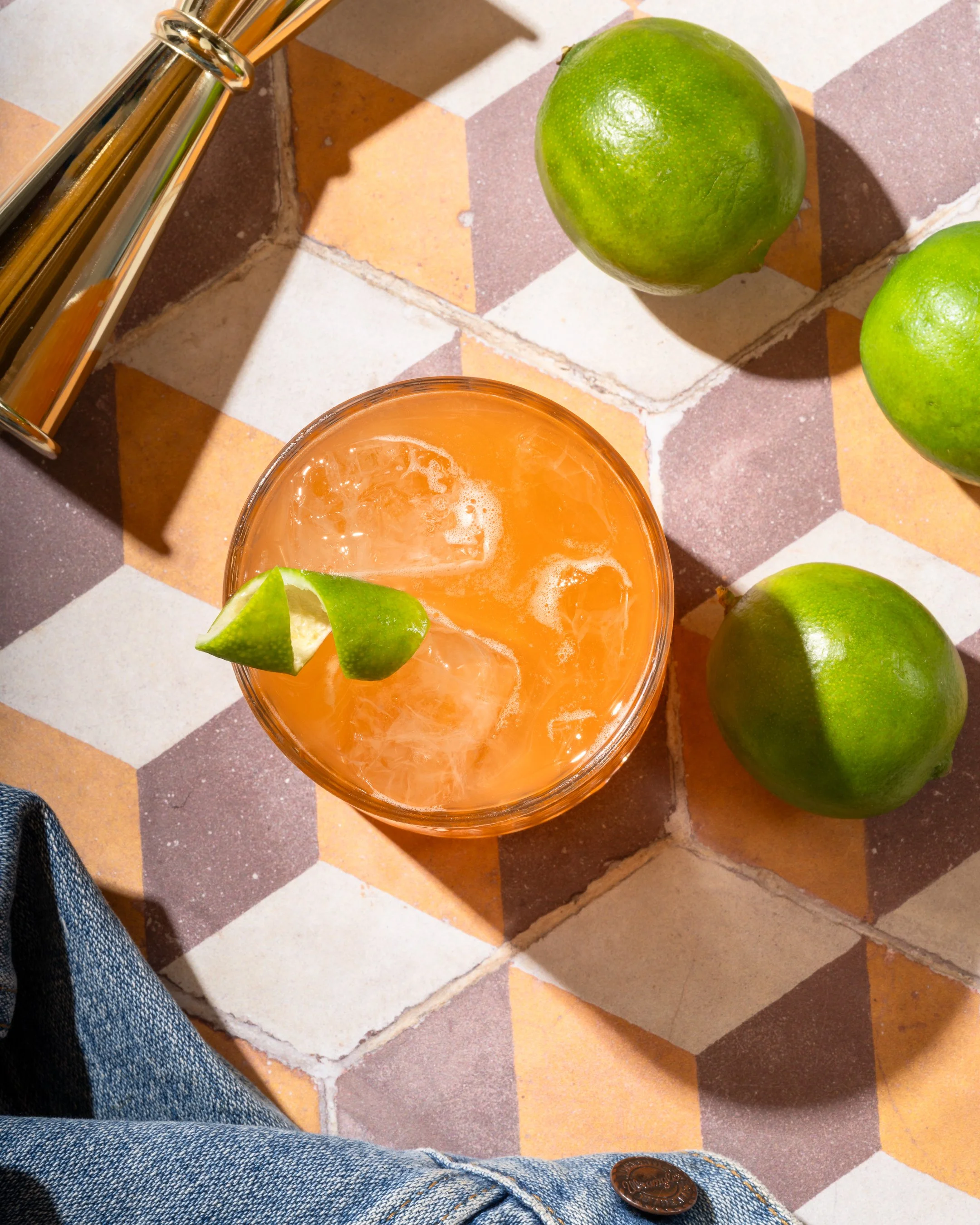 A glass of orange drink with ice and lime wedge on a colorful tiled table surrounded by whole limes and a lemon squeezer.