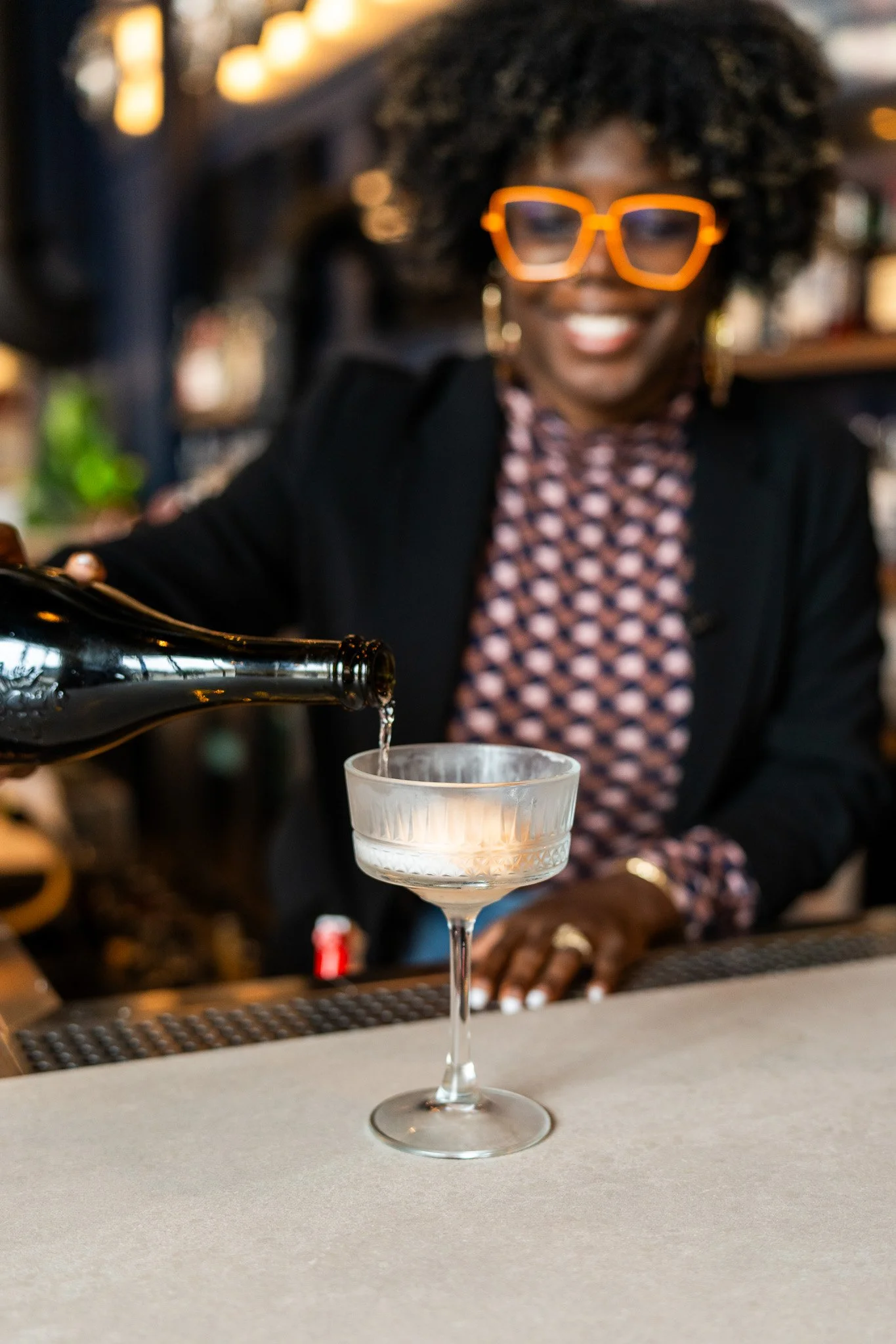 Woman with curly hair and orange glasses pouring a drink into a cocktail glass at a bar.