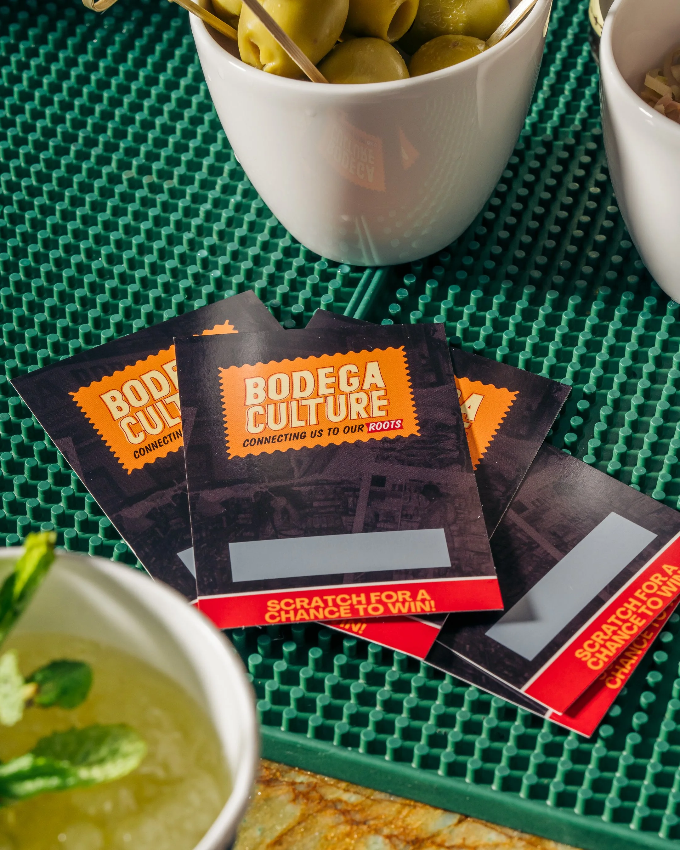 Brochures for Bodega Culture on a green textured table with bowls of olives and a drink.