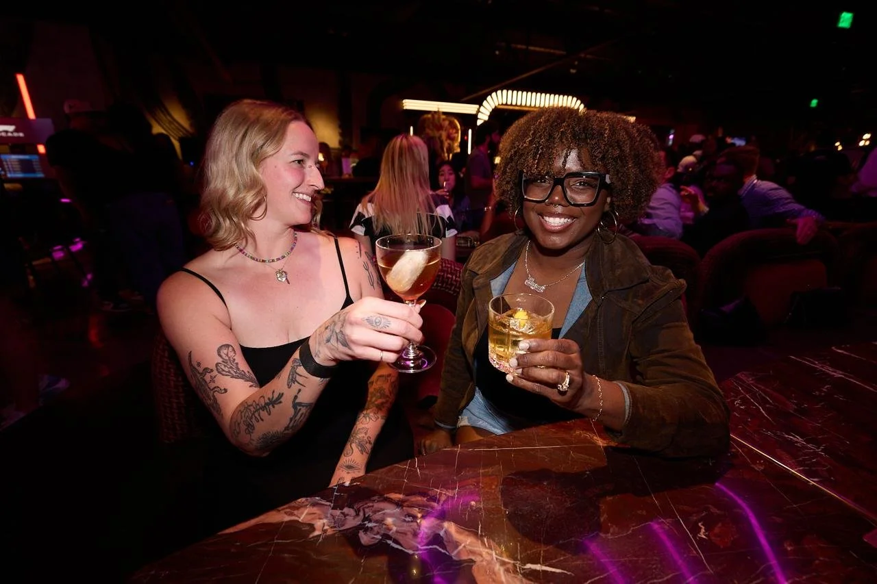 Two women sitting at a bar or nightclub, smiling and holding drinks, surrounded by other patrons in a lively, colorful setting.