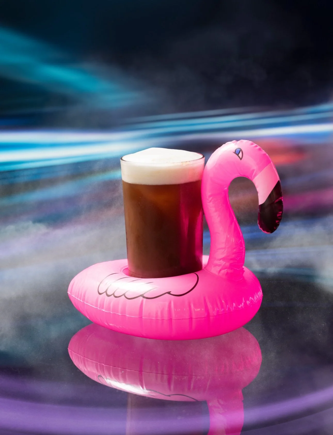 A pink inflatable flamingo drink holder with a glass of dark beer inside it, on a reflective surface with a colorful blurred background.