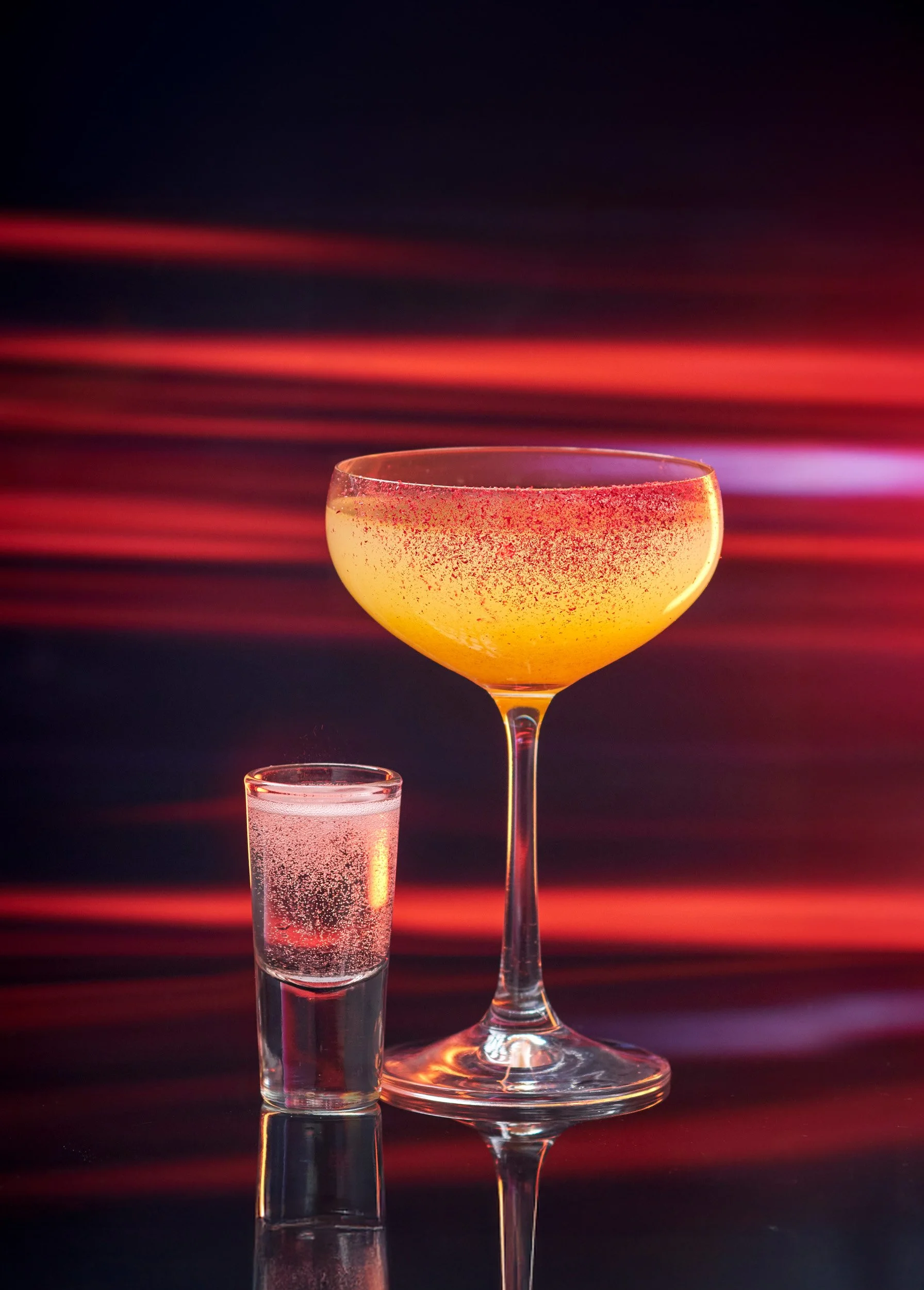 A large yellow cocktail in a glass with a red rim and a smaller clear glass of water with bubbles, set against a dark background with red and purple streaks of light.