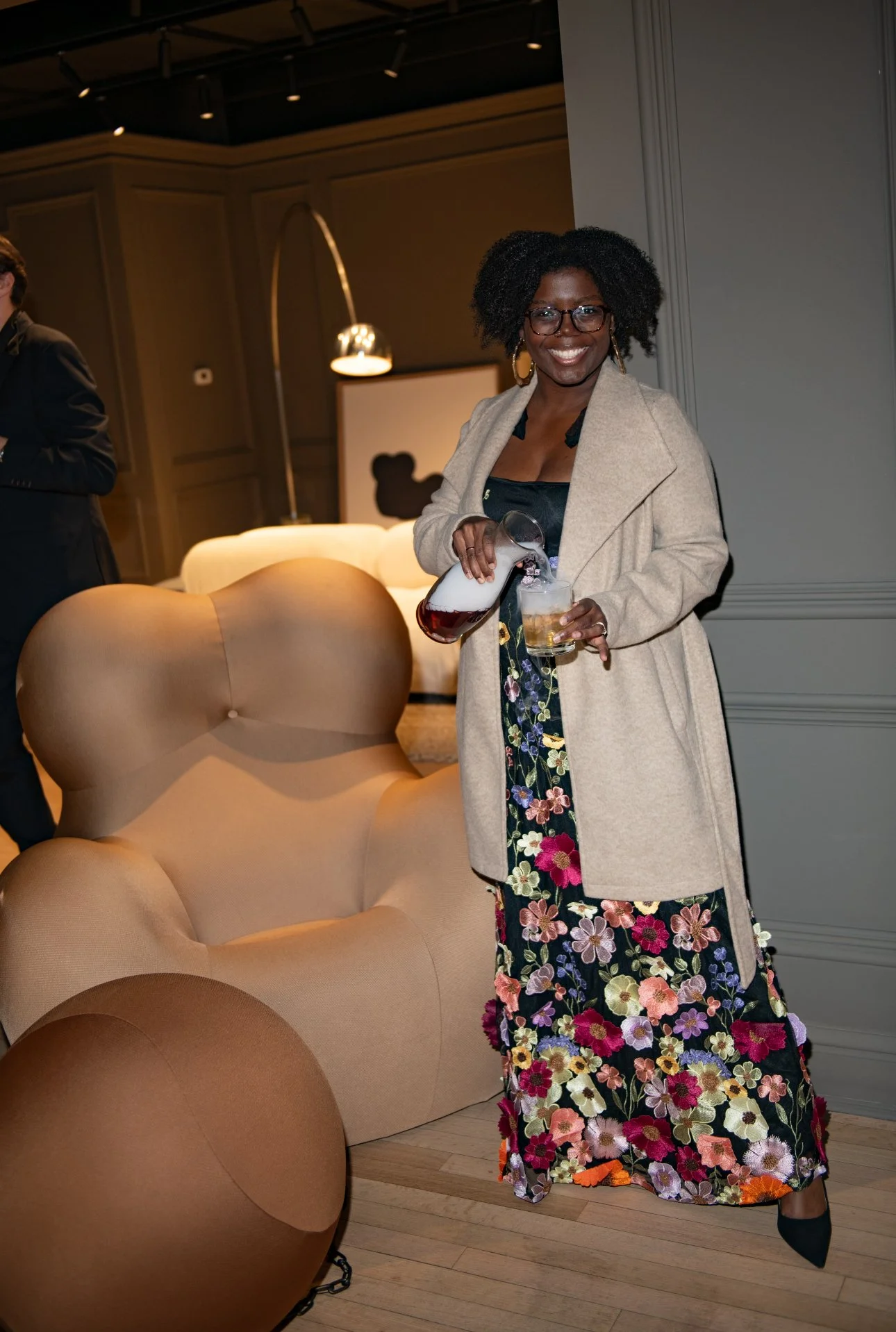 A woman with glasses and curly black hair is pouring a drink with foam into a glass, standing next to beige and brown chairs in a dimly lit, stylish interior.