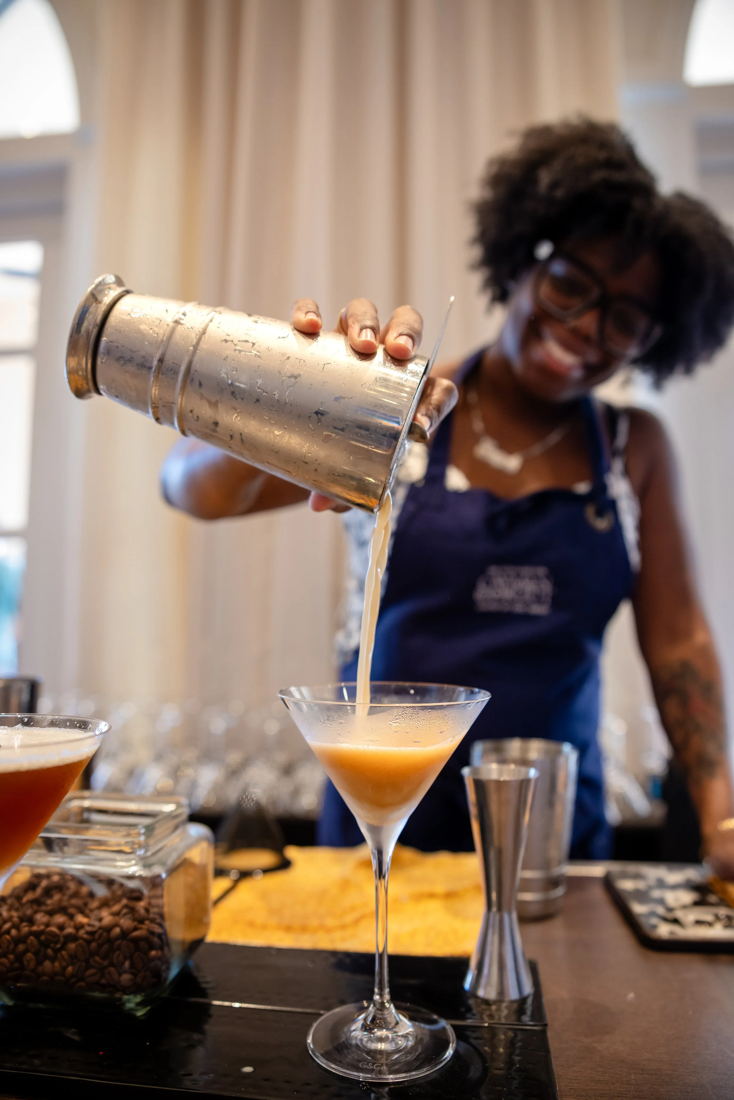 A woman pouring a creamy cocktail from a cocktail shaker into a martini glass, with coffee beans and other bar tools on the table.
