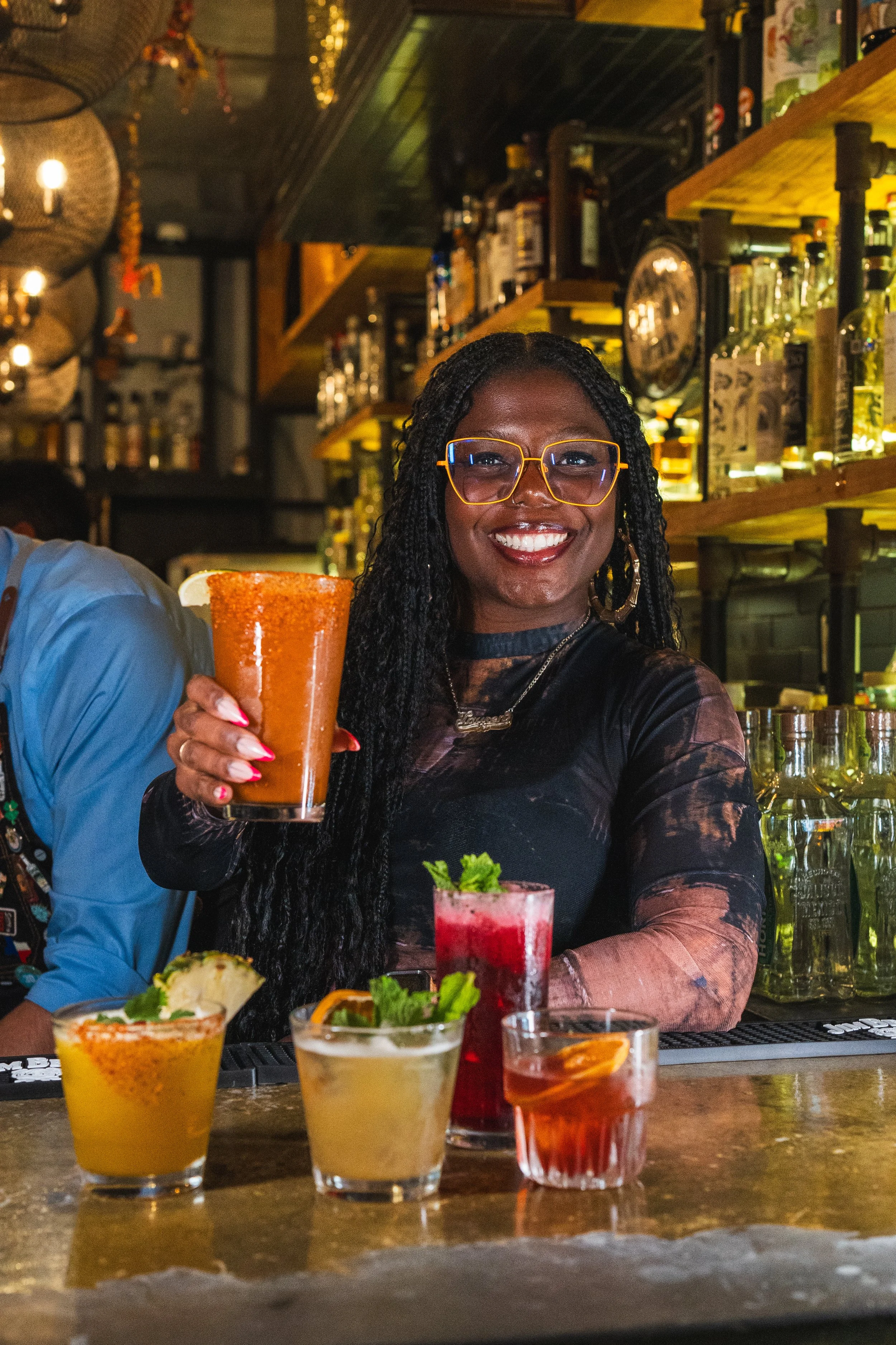 A woman with glasses and long black braids smiling and holding a cocktail at a bar, with various drinks on the bar counter and bottles of alcohol in the background.