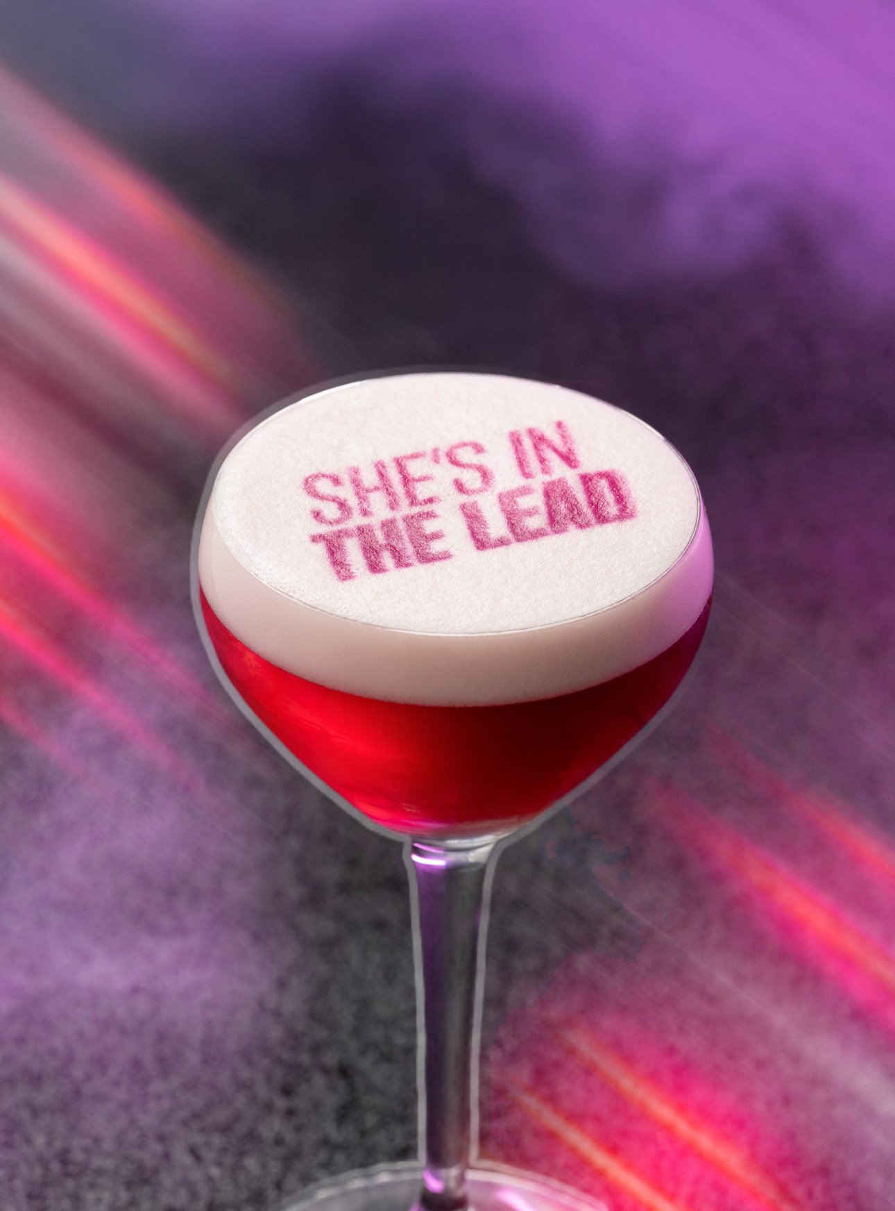 A cocktail glass with red liquid and a white foam top that has pink text reading "SHE'S IN THE LEAD". The background is blurred with purple and pink lighting.