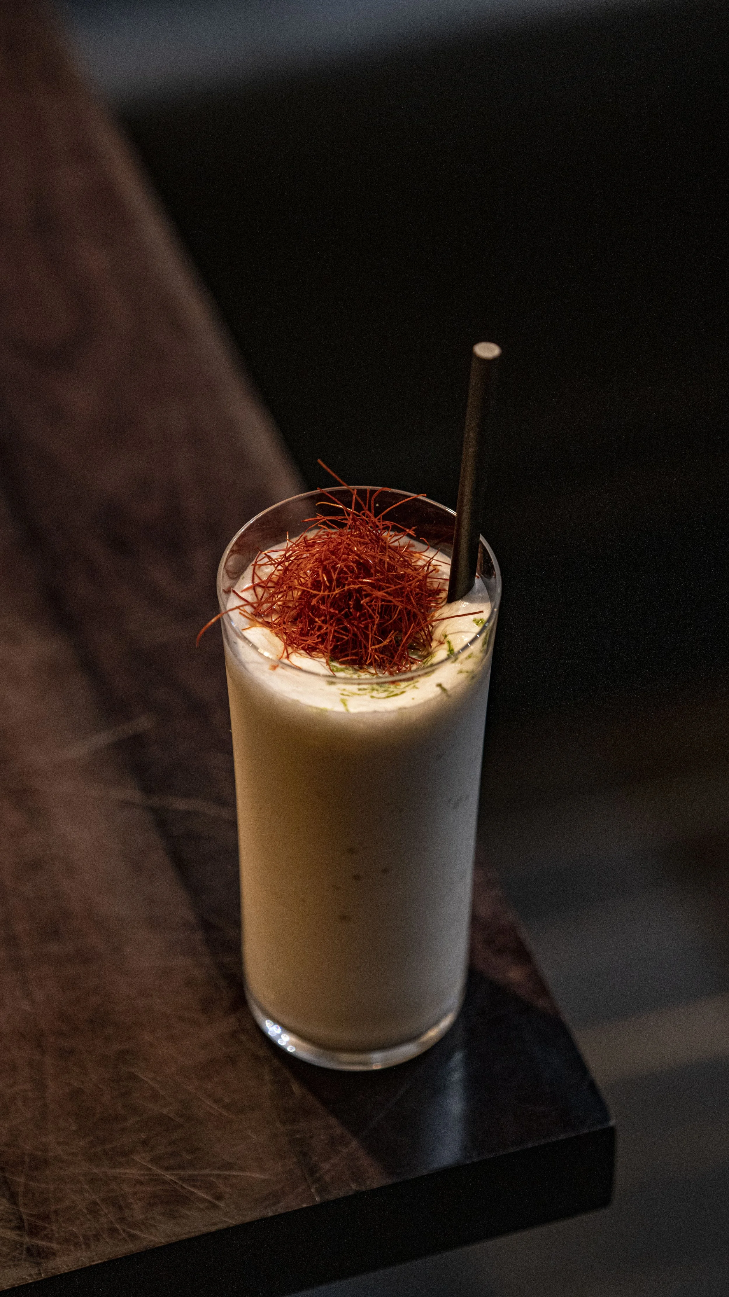 Tall glass of creamy cocktail garnished with red herbs and a black straw, placed on a dark wooden surface.