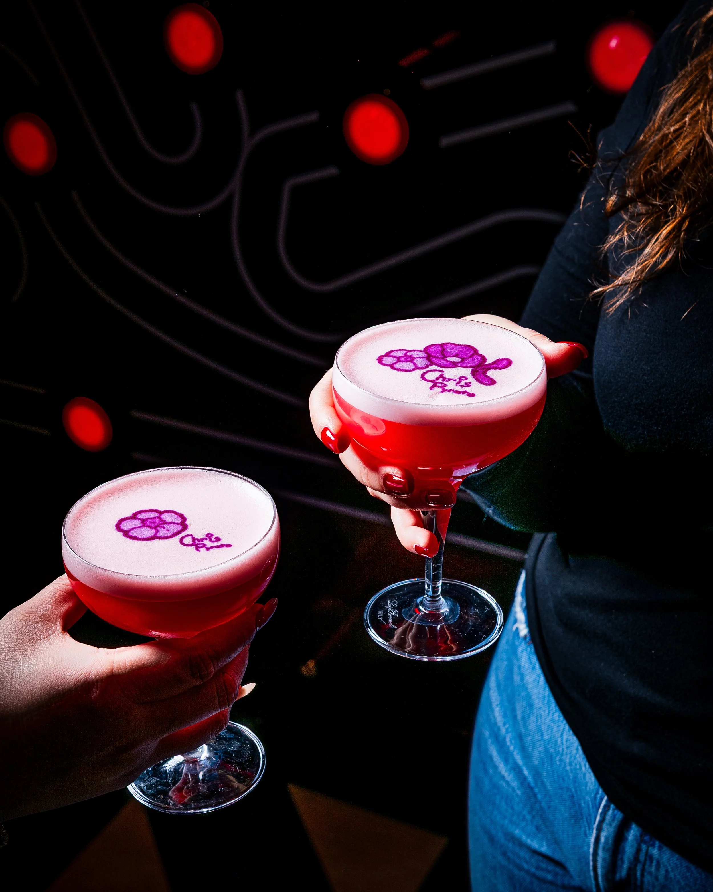 Two women holding pink drinks in coupe glasses with a purple logo on top, dark background with abstract red lights.