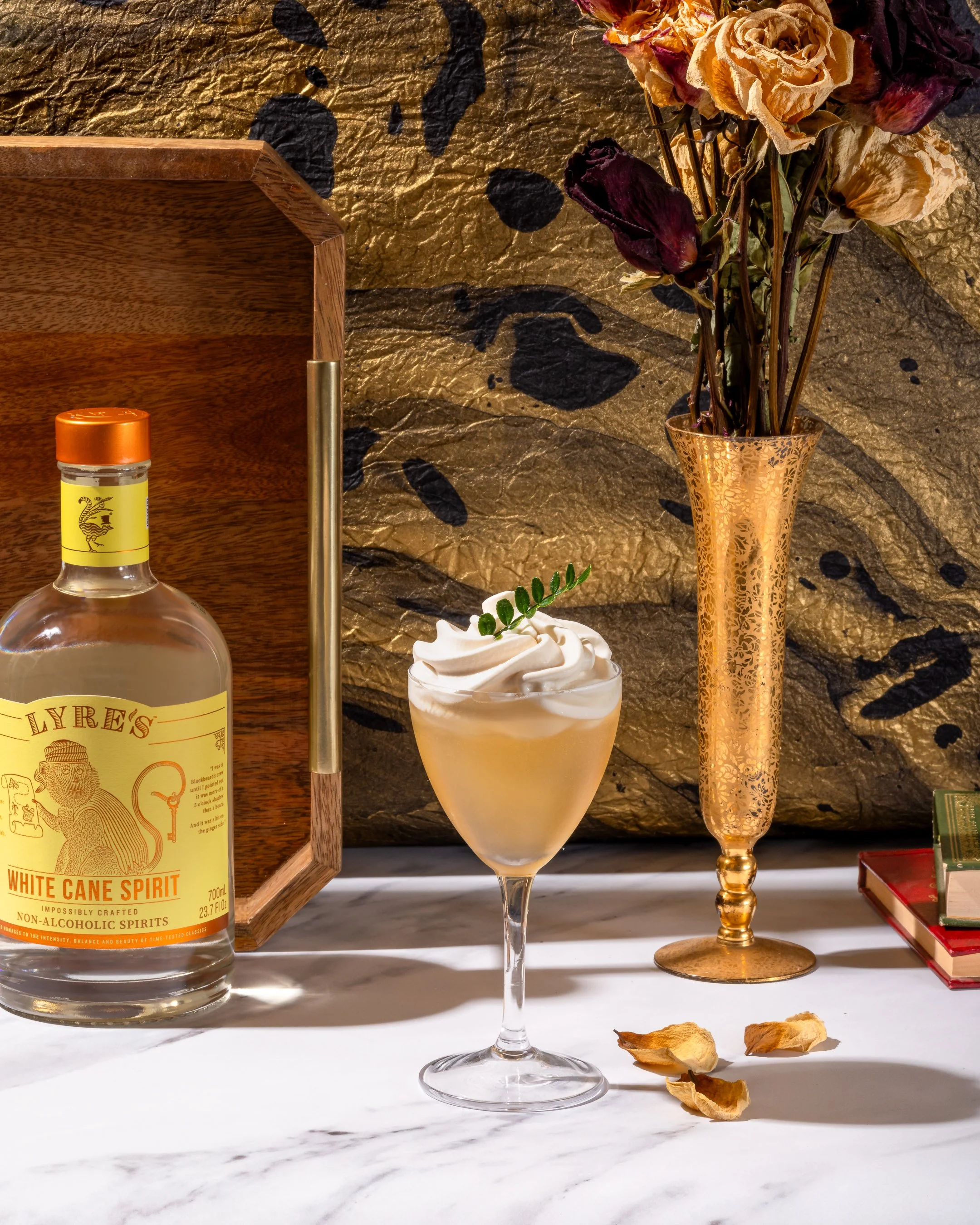 A cocktail in a stemmed glass topped with whipped cream and a small green leafy garnish, a bottle of non-alcoholic white cane spirit, a tall gold-patterned vase with dried roses, a small green plant, some dried petals, and a stack of books on a white