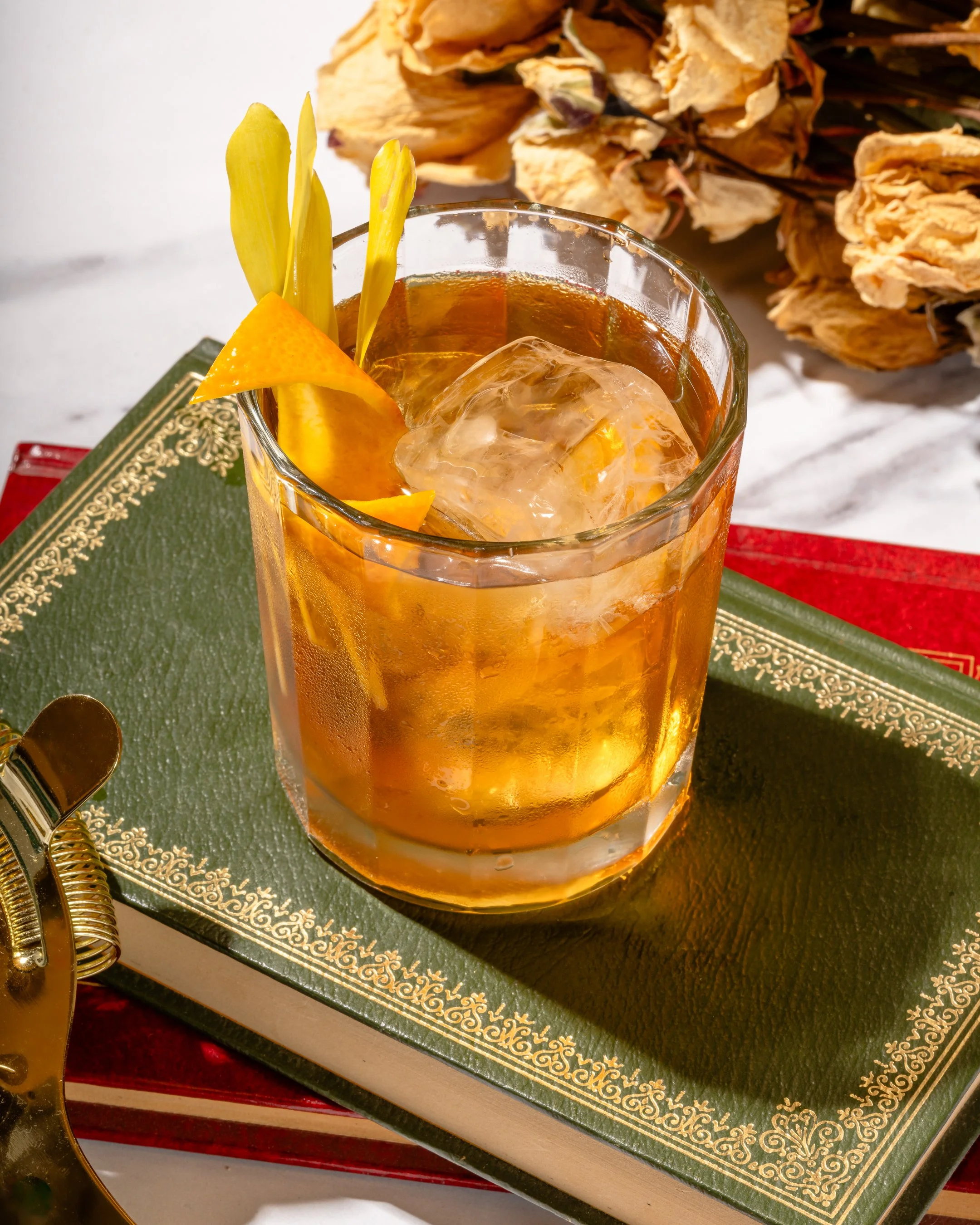 A glass of iced amber-colored cocktail garnished with lemon and orange slices, placed on a decorative green book with a red book underneath, with dried flowers in the background.