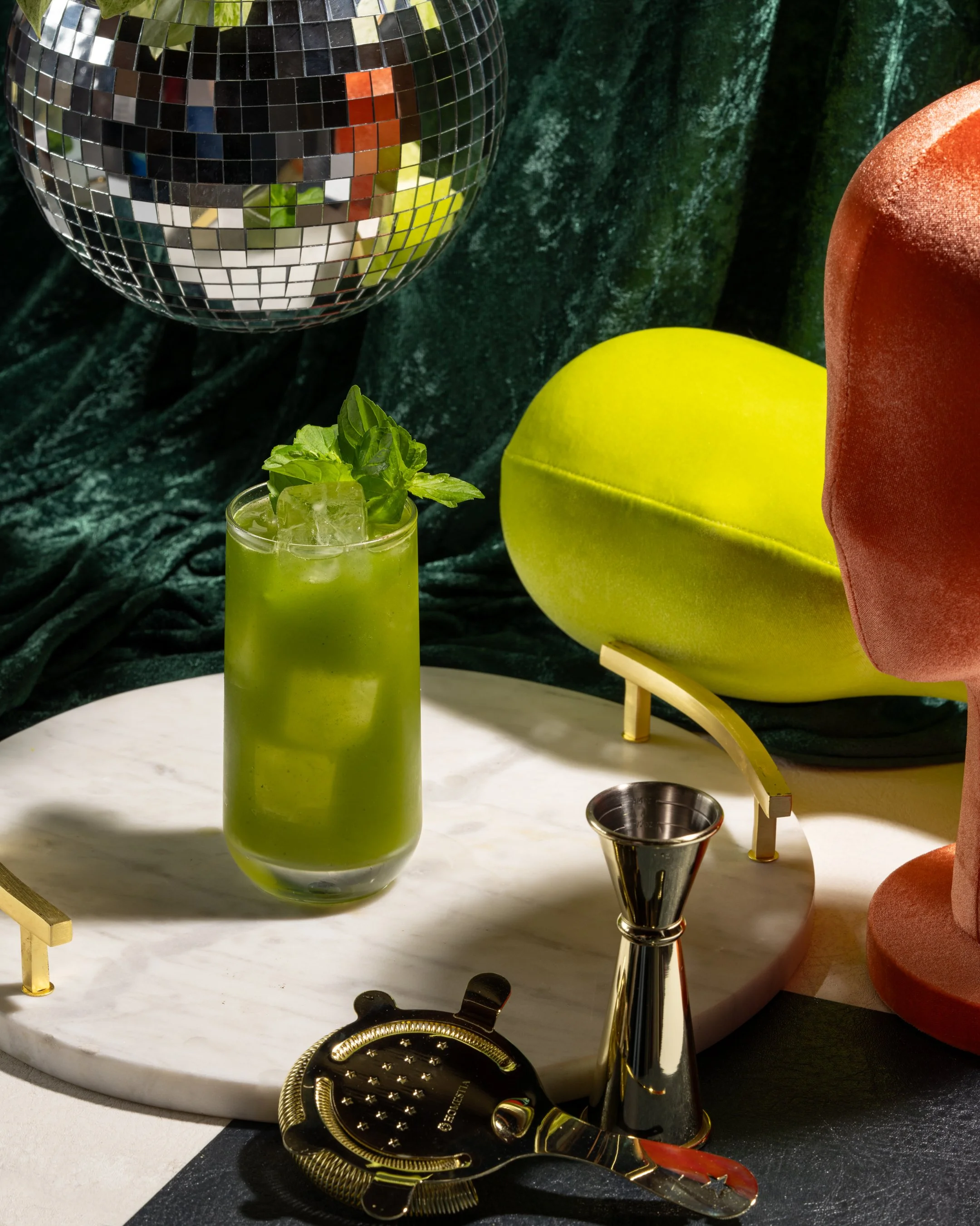A green cocktail with ice and basil on a white marble surface, with a disco ball hanging above, green and red velvet fabrics in the background, and bar tools nearby.