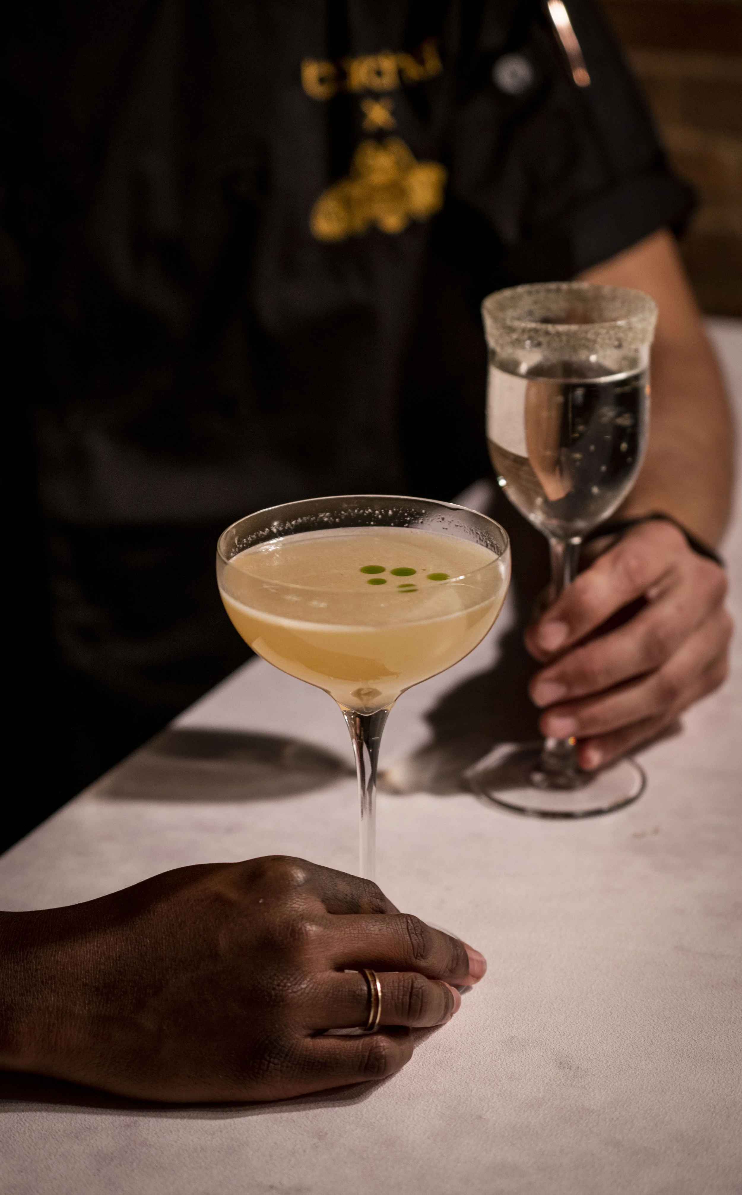Two people holding cocktails, one in a coupe glass with a light yellow drink garnished with green dots, and the other holding a champagne flute with clear sparkling wine. The scene appears to be at a bar or restaurant.