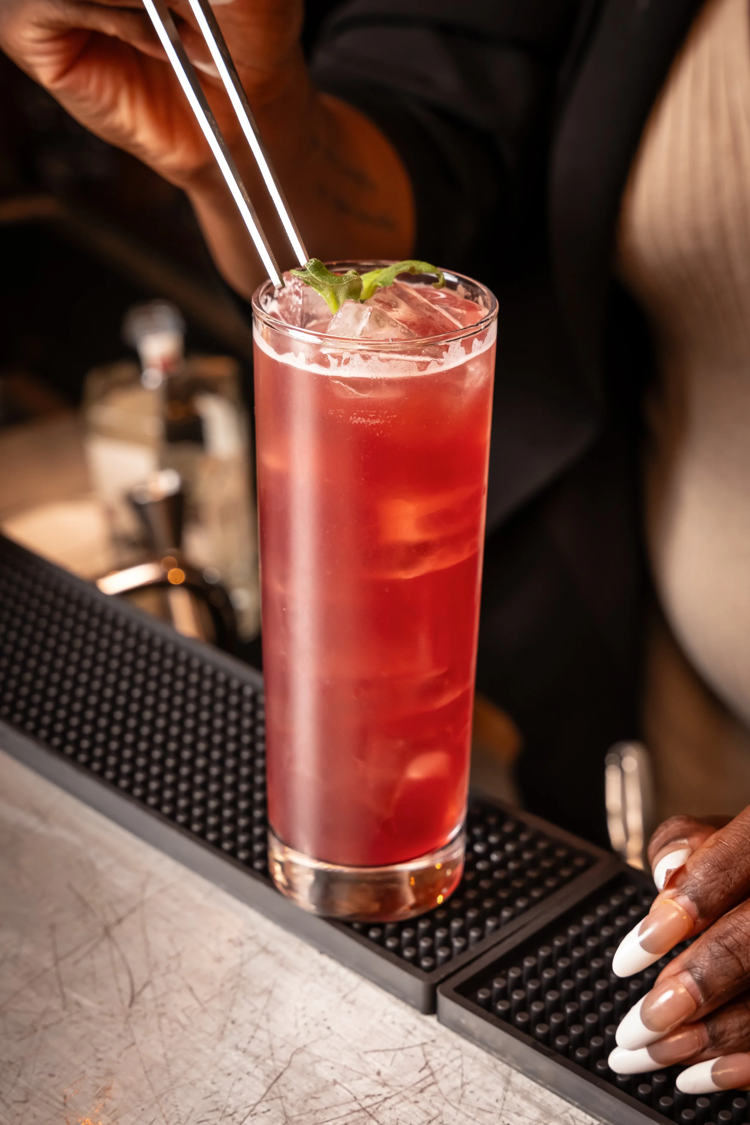 Tall glass of pink cocktail with ice, garnished with a lime wedge and a sprig of mint, on a bar counter.