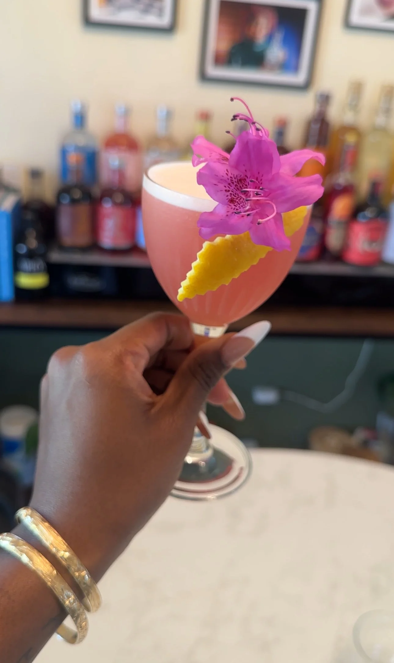 A person holding a pink cocktail garnished with a pink flower and a yellow lemon twist. The background features bottles on a bar shelf.