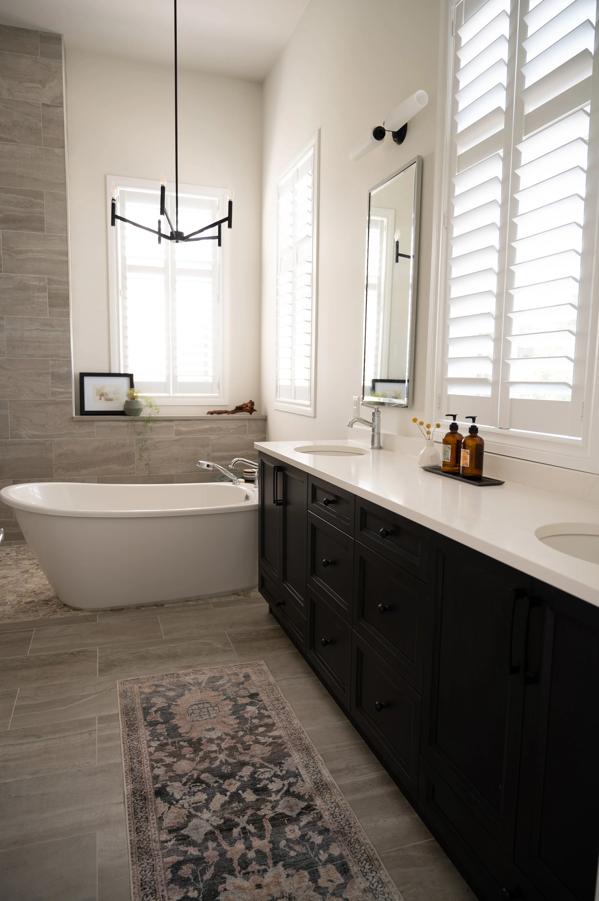Modern bathroom with a white bathtub, black vanity with double sinks, large window with shutters, decorative rug, and contemporary pendant light fixture.