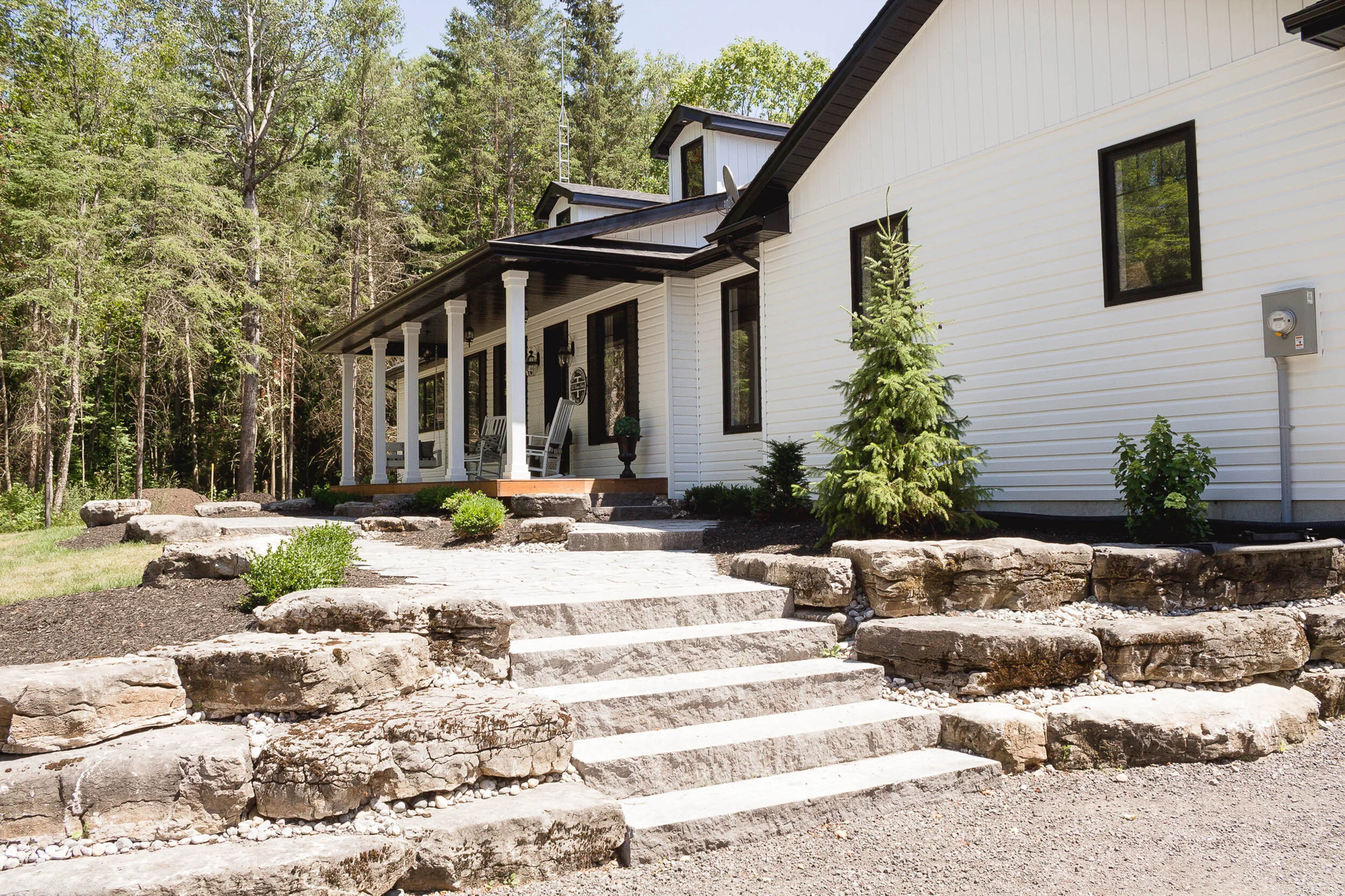 Front view of a white house with a porch and steps, surrounded by a landscaped yard with rocks and small trees, located in a wooded area.