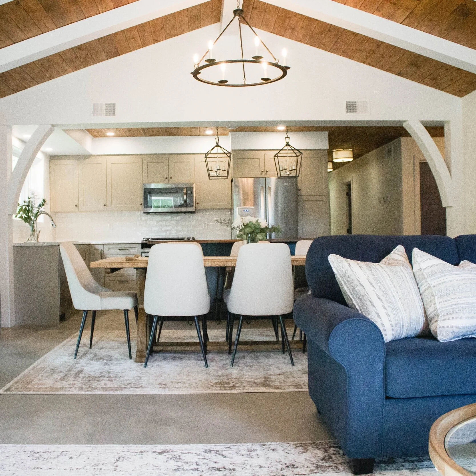 Open-concept living and dining area with a kitchen in the background, a navy blue sofa with pillows in the foreground, and a wooden dining table with white chairs. Chandeliers hang from the wooden vaulted ceiling.