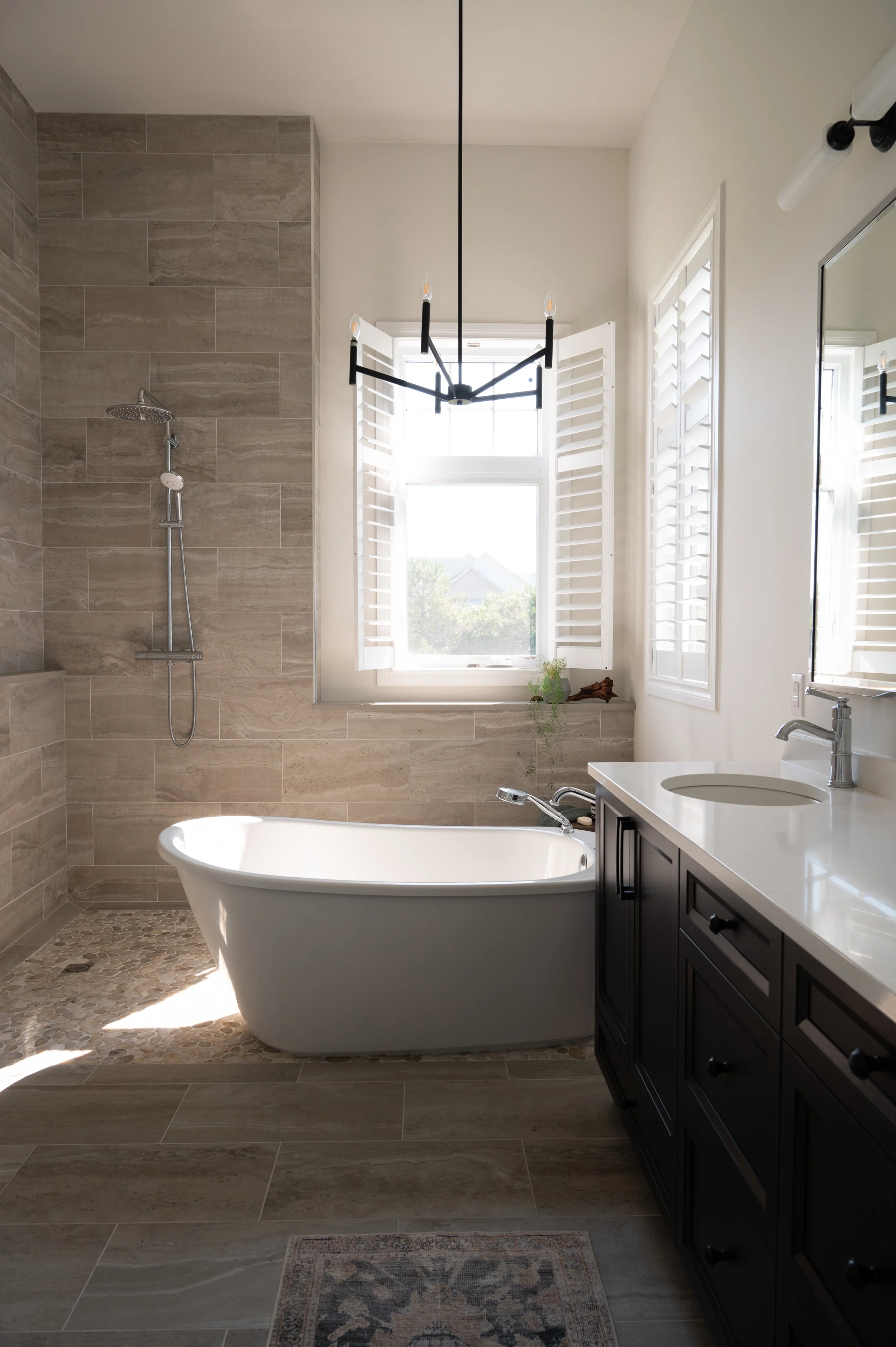 Modern bathroom with a freestanding bathtub, a walk-in shower, dark wood vanity with a white countertop, and windows with white shutters, illuminated by a contemporary black chandelier.