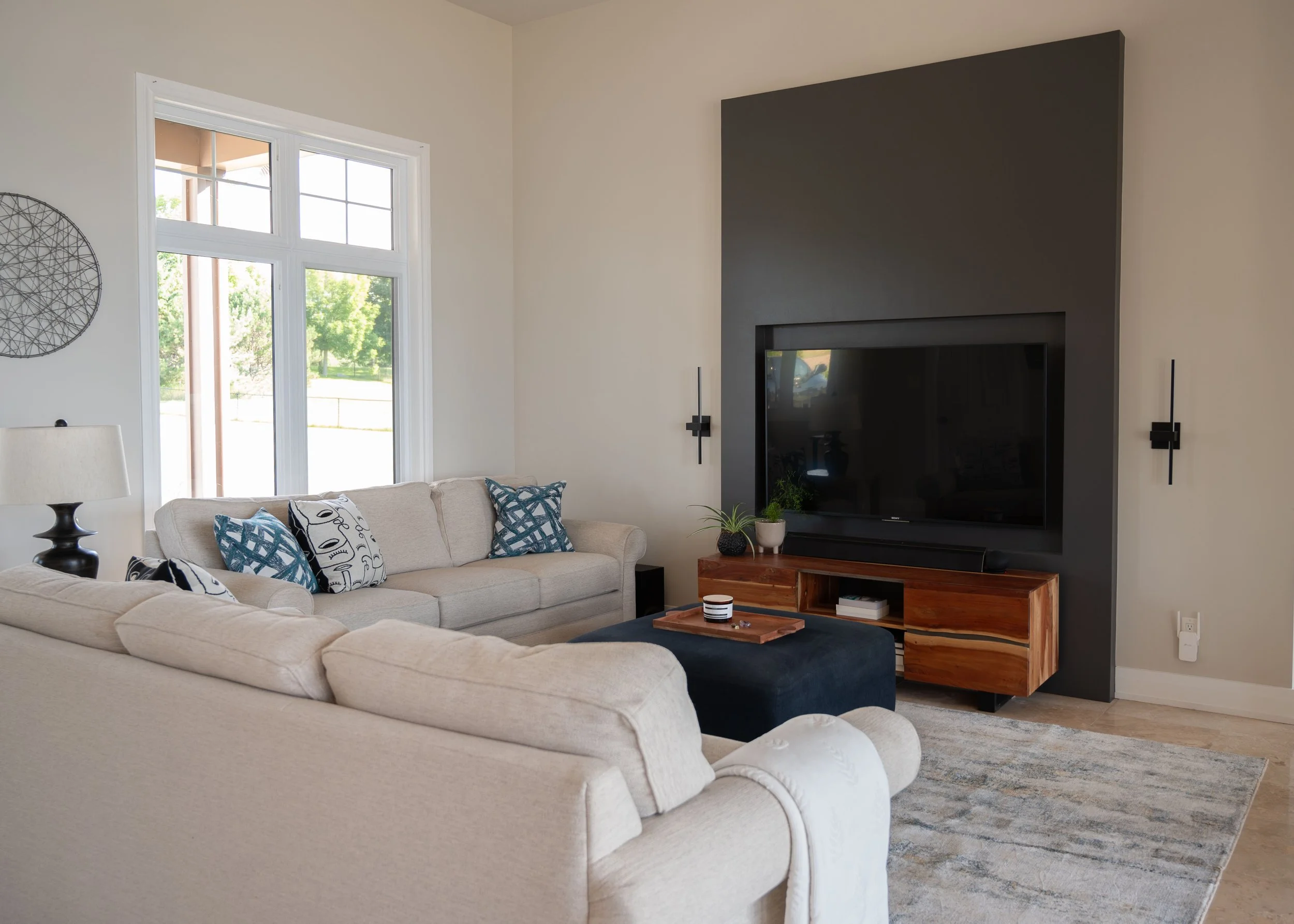 Living room with beige sectional sofas, blue and patterned throw pillows, a large window, a black accent wall with a mounted TV, a wooden media console, potted plants, a beige rug, and side wall sconces.