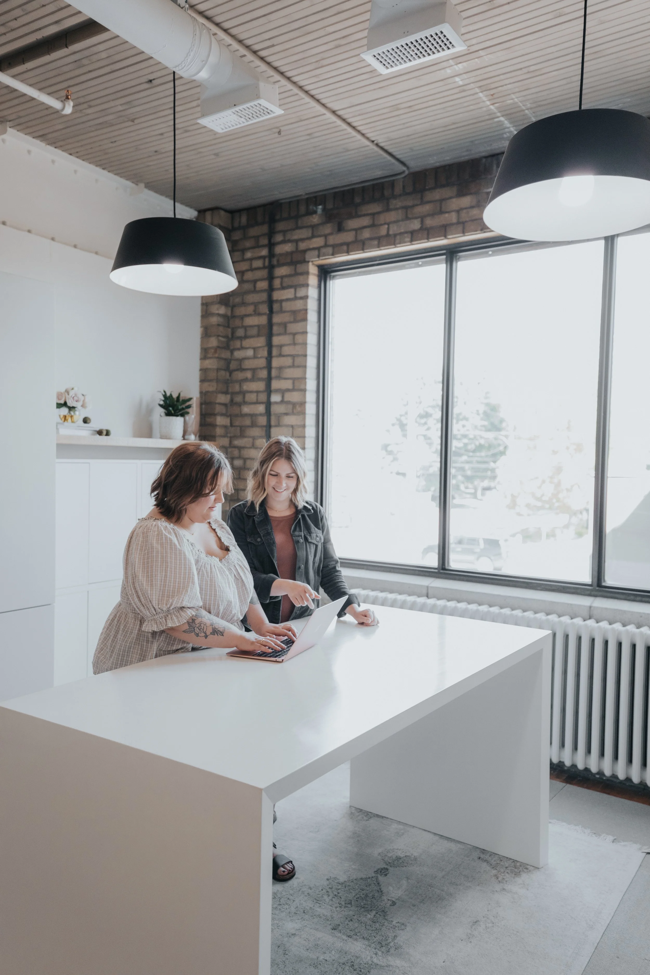 Two women collaborating at a white table in a modern office with large windows, exposed brick wall, ceiling lights, and minimal decor.