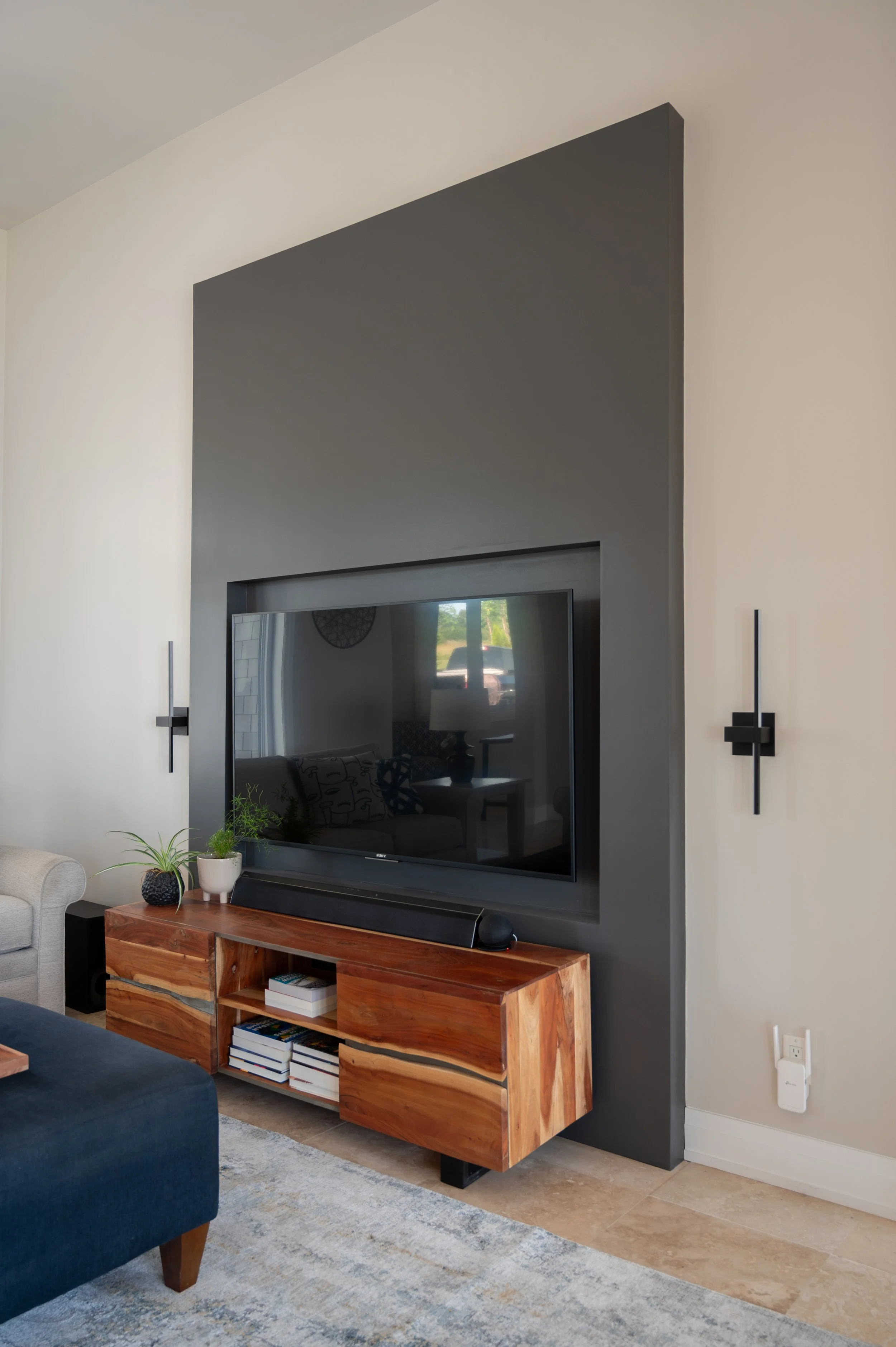 Living room with wall-mounted TV, black and gray wall, wooden media console, beige and blue furniture, green plants on the console, and decorative sconces on the wall.