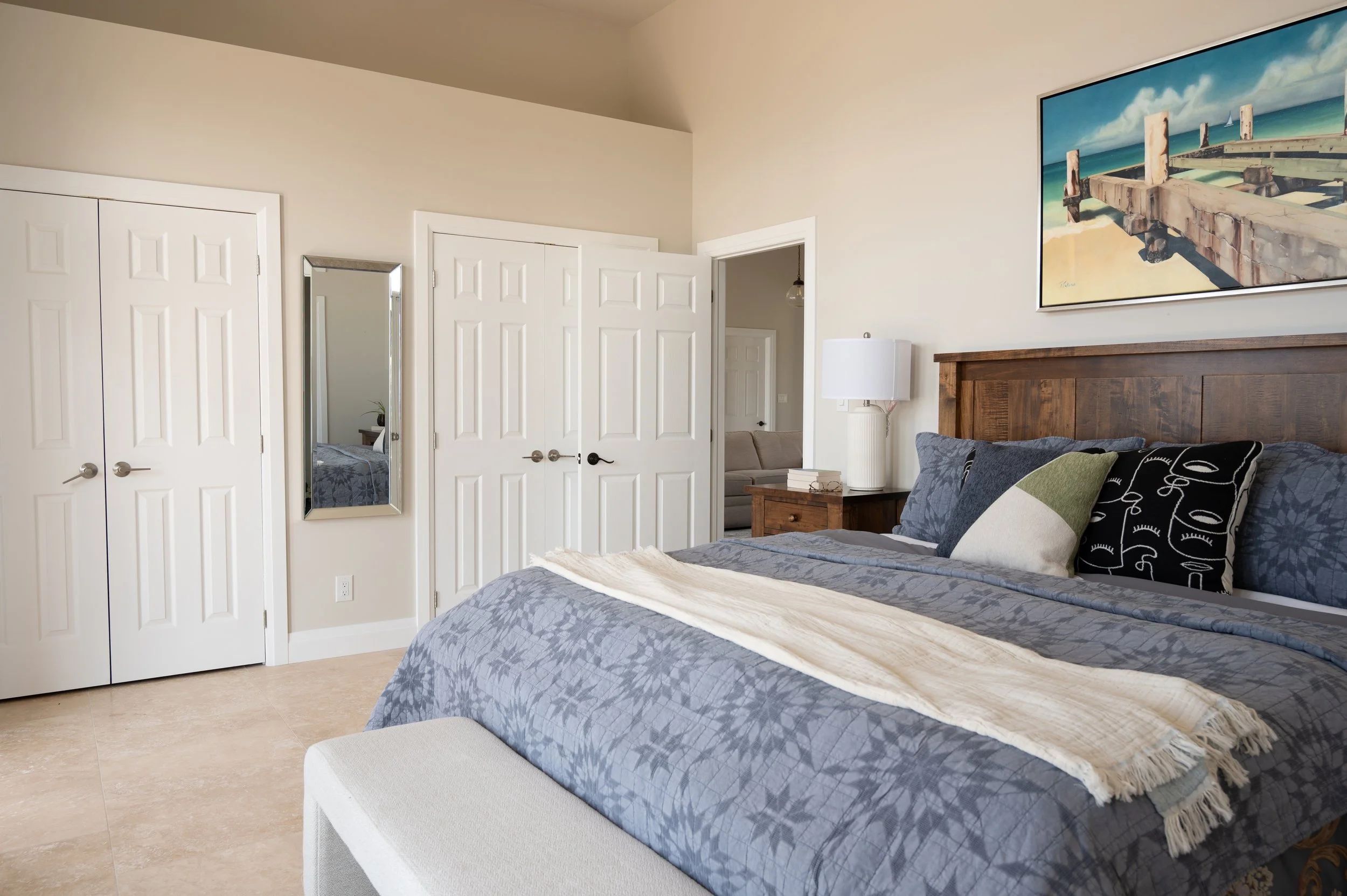 A bedroom with a bed, pillow, beige blanket, wooden headboard, white lamp, painting of a pier, mirror, and a closet.